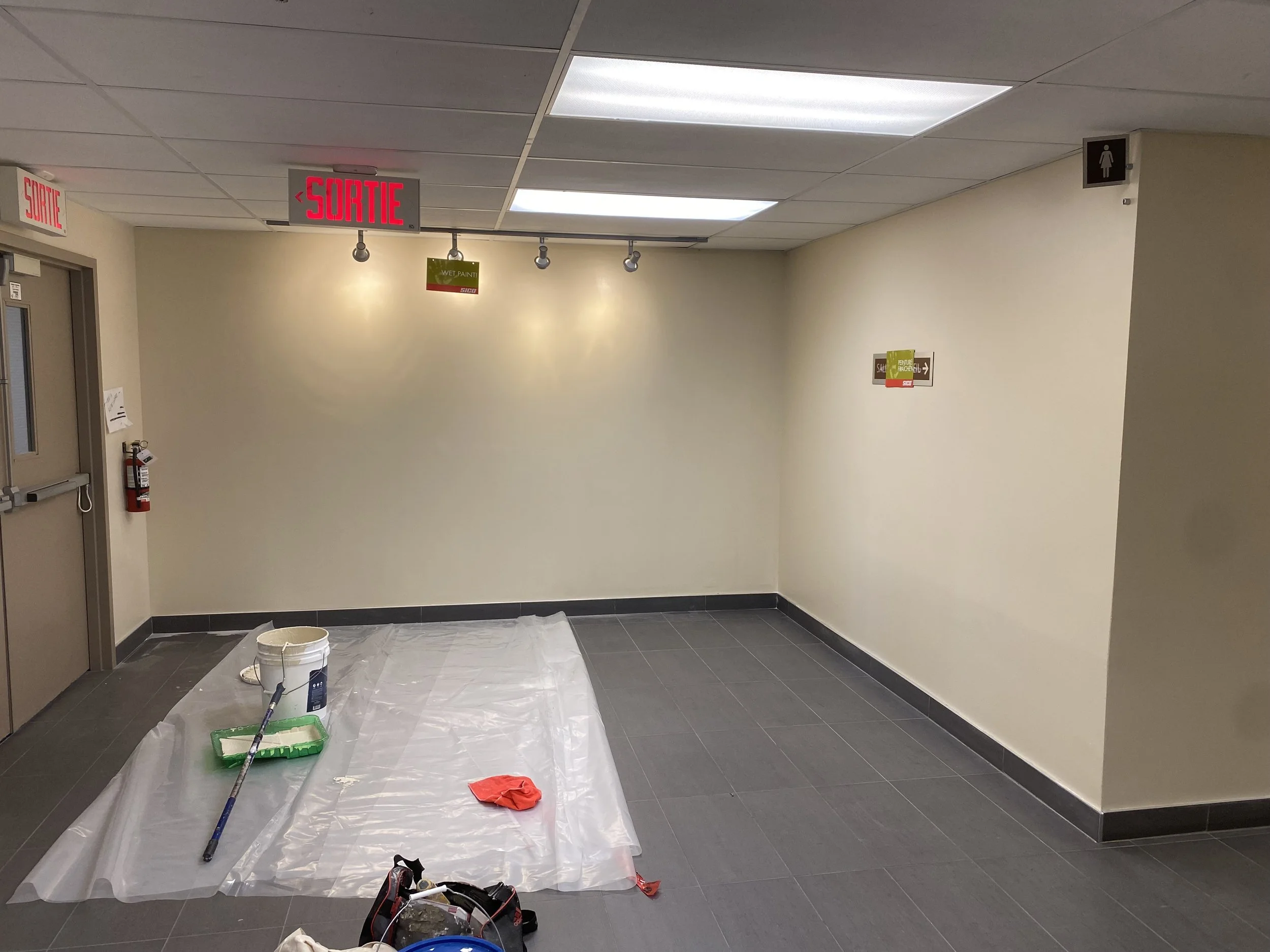 An indoor hallway with ceiling lights, grey tile floor, and beige walls. A painting or sign with the text 'Wet Paint' hangs on the wall. A white sheet covers the floor with painting supplies, including a paint roller in a green tray, a paint bucket, 