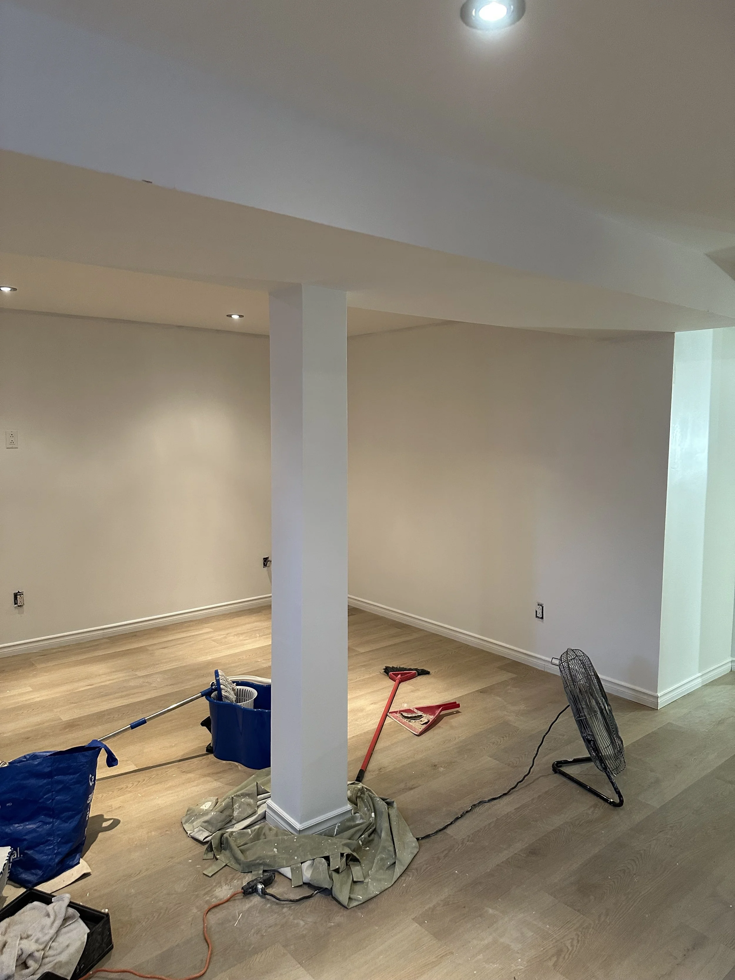Interior room under renovation with tools on the floor, including a red trowel, black fan, and blue bucket, and scattered materials.