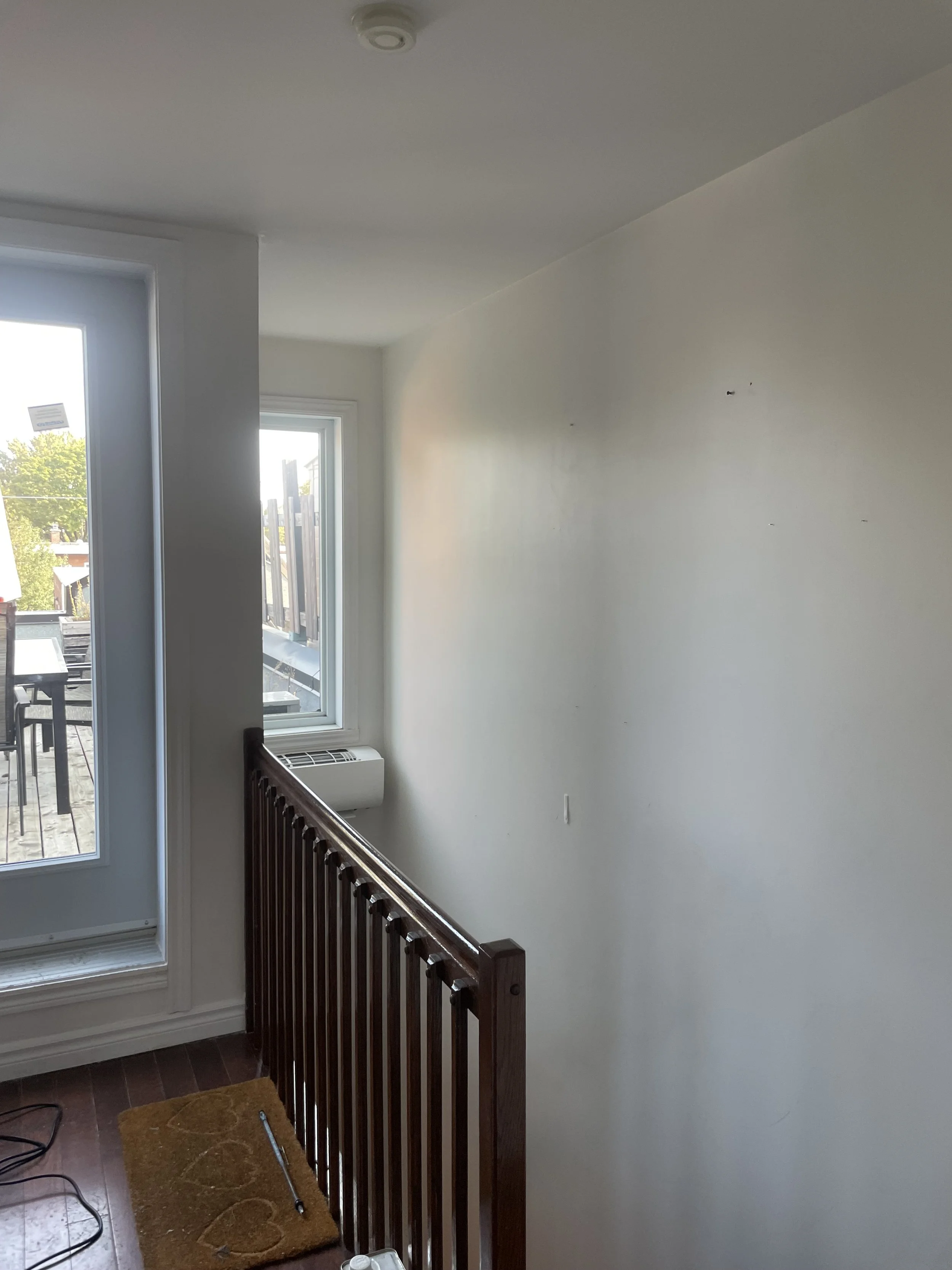 An interior of a room with a white wall, a window, a dark brown wooden railing, and a small brown rug on wooden floor. A tool is resting on the rug and an air vent is below the window.