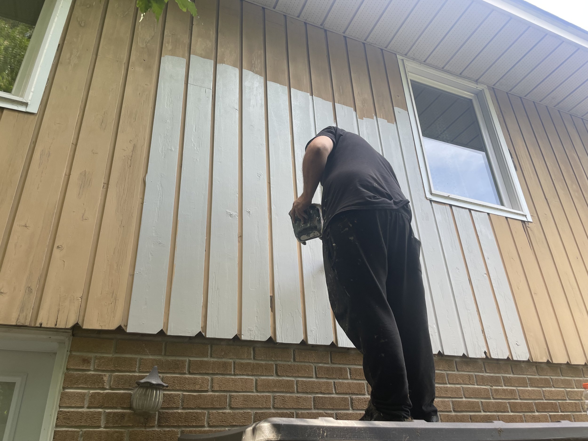 Person painting the exterior wall of a house beige, with a paintbrush in hand, standing on a small step ladder. The house has horizontal wooden siding and a brick foundation. There is a window and a outdoor wall light fixture on the house.