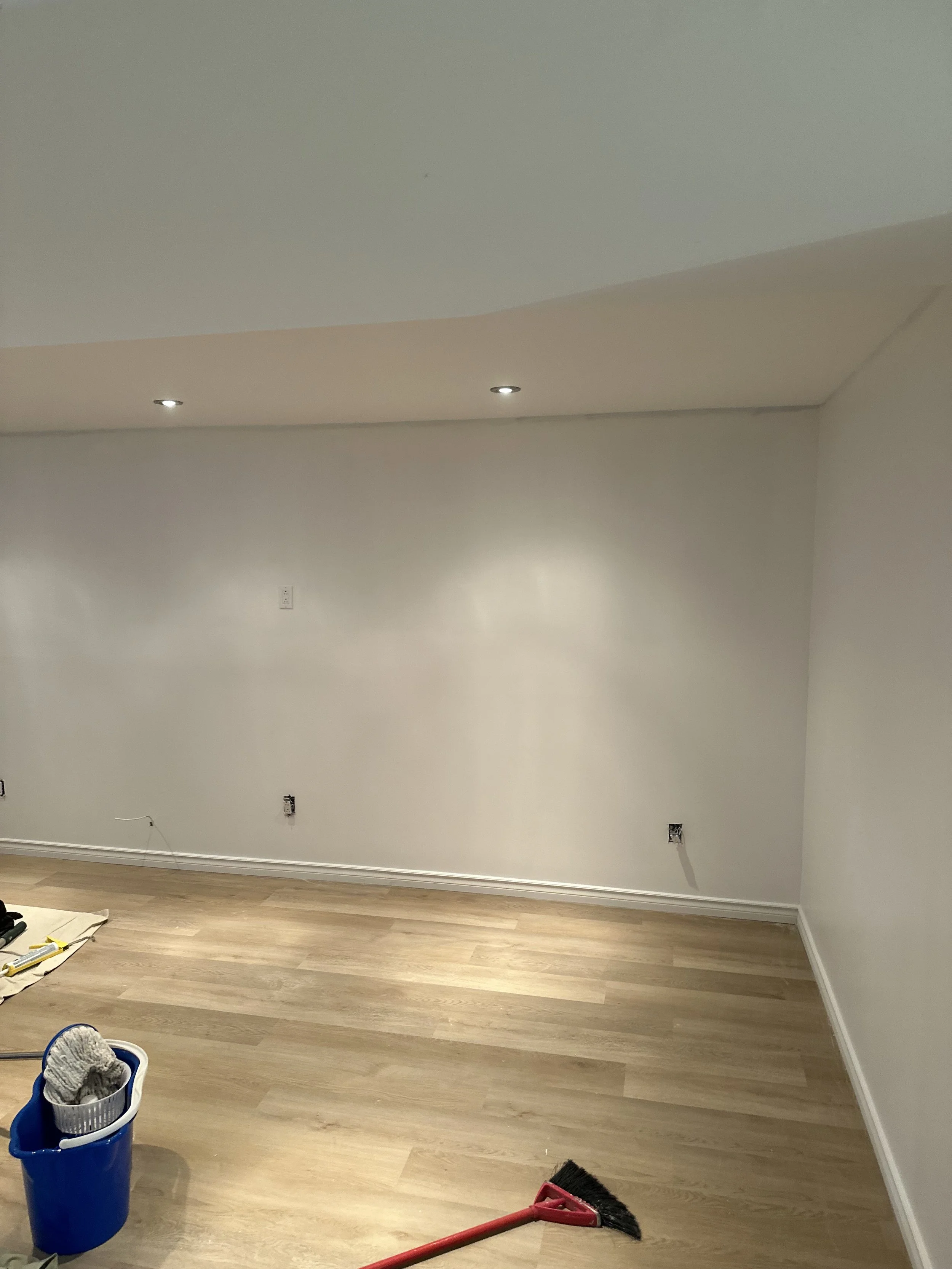 A partially renovated room with beige wooden flooring and white walls. Several electrical outlets are visible on the wall. Cleaning tools, including a broom with a red handle and a blue bucket with cleaning supplies, are on the floor. Recessed lighti