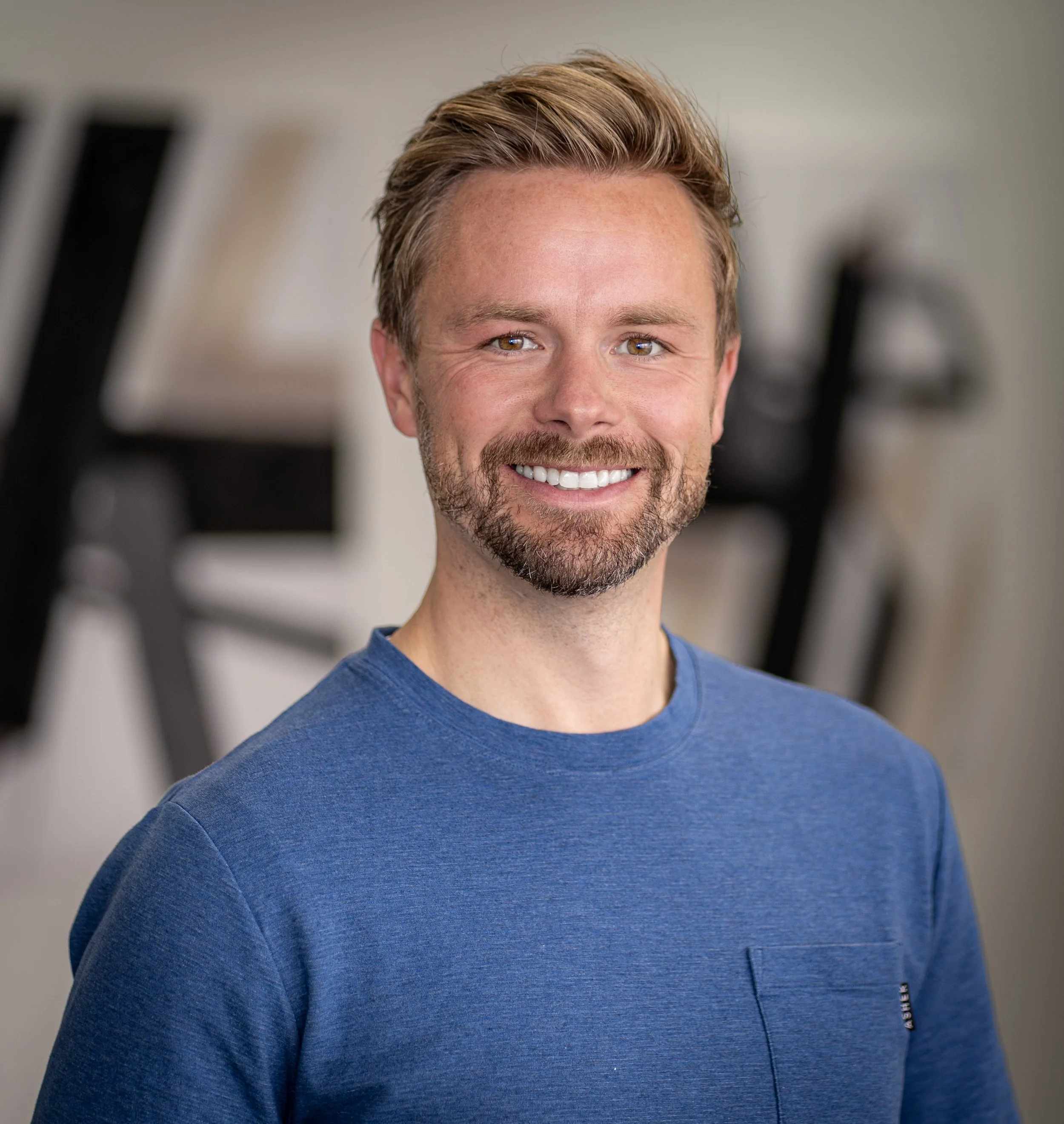 Close-up of a smiling man with brown hair, beard, and blue shirt, indoors.