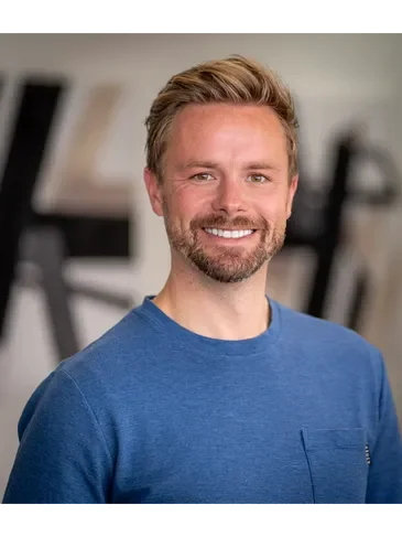 Portrait of a man with blonde hair and a beard, smiling, wearing a blue t-shirt, in an indoor setting.