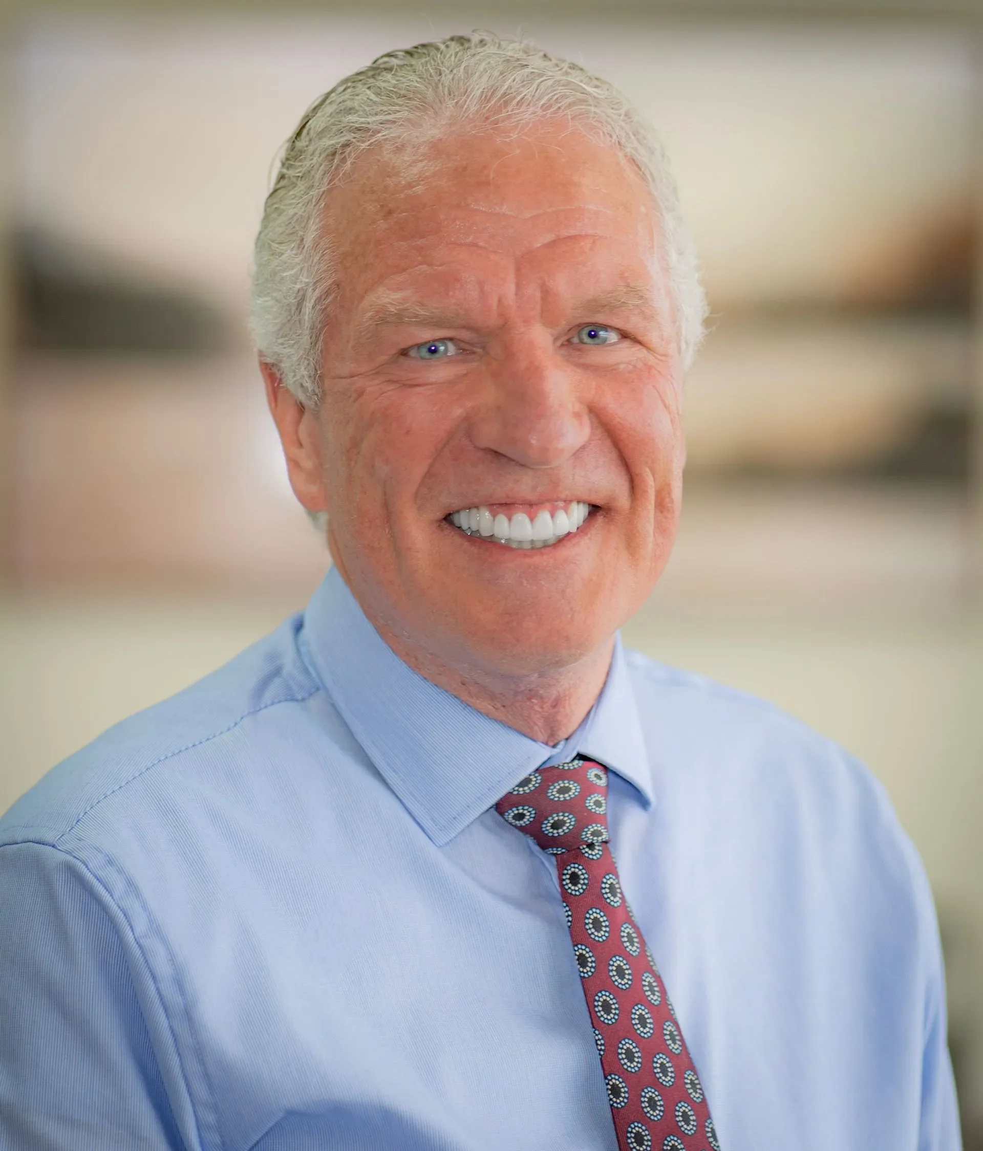 A smiling elderly man with white hair, dressed in a light blue shirt and a patterned red tie, standing outdoors with a blurred background.