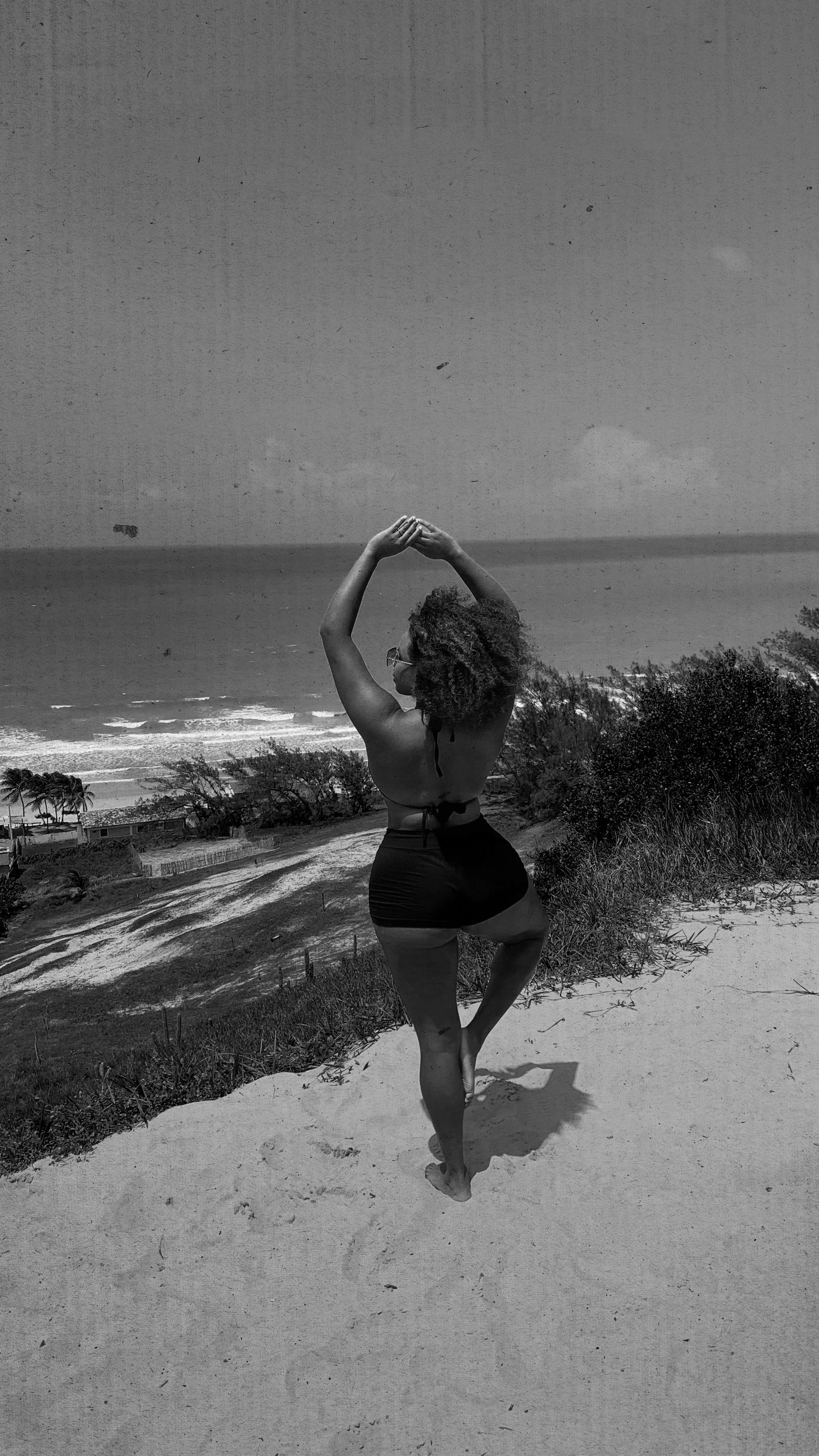 A woman practicing yoga on a beach at sunset, wearing a sports bra and shorts, with her arms raised above her head in a pose.