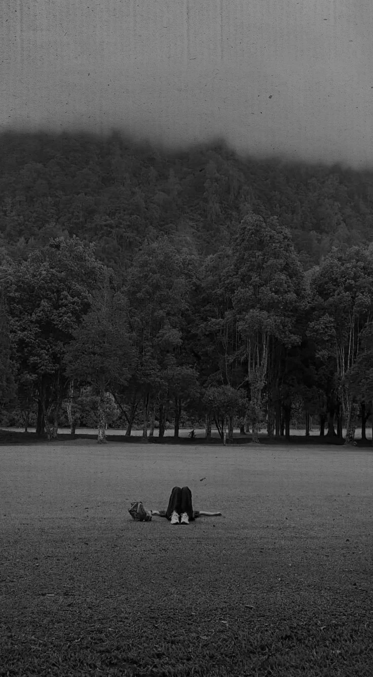 A person sitting on the grass with their head bent down, near a backpack, in a park with a dense tree line and mountains in the background. The photo is in black and white.