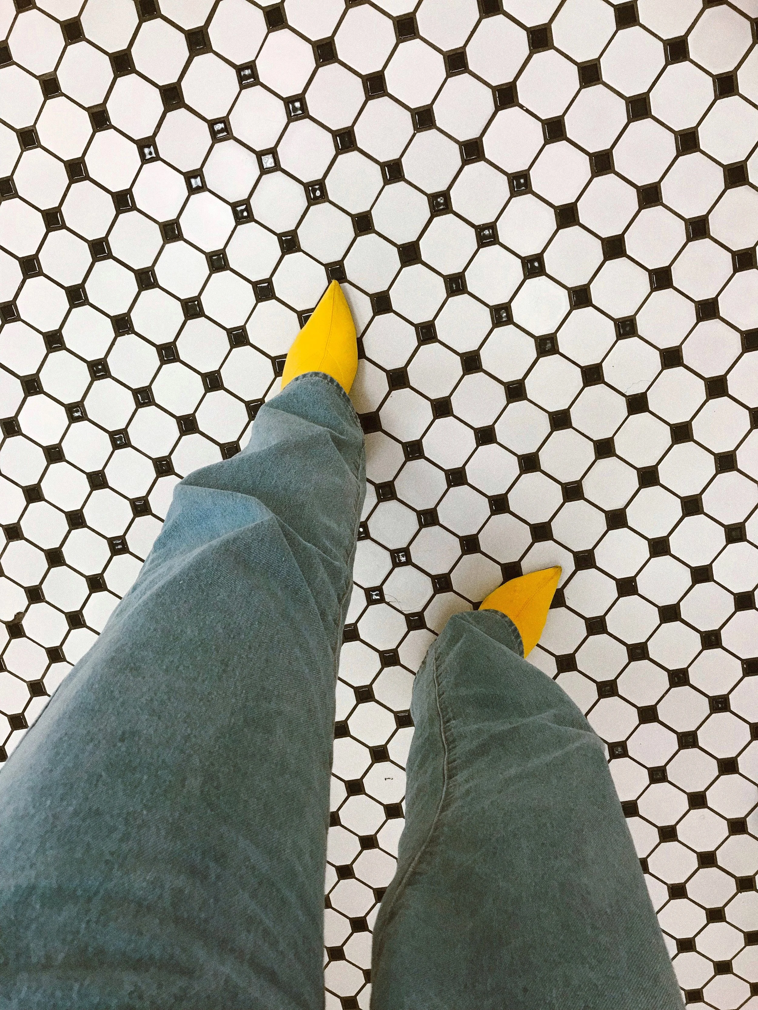 A person's legs in blue jeans and yellow shoes walking on a white and black patterned tile floor.