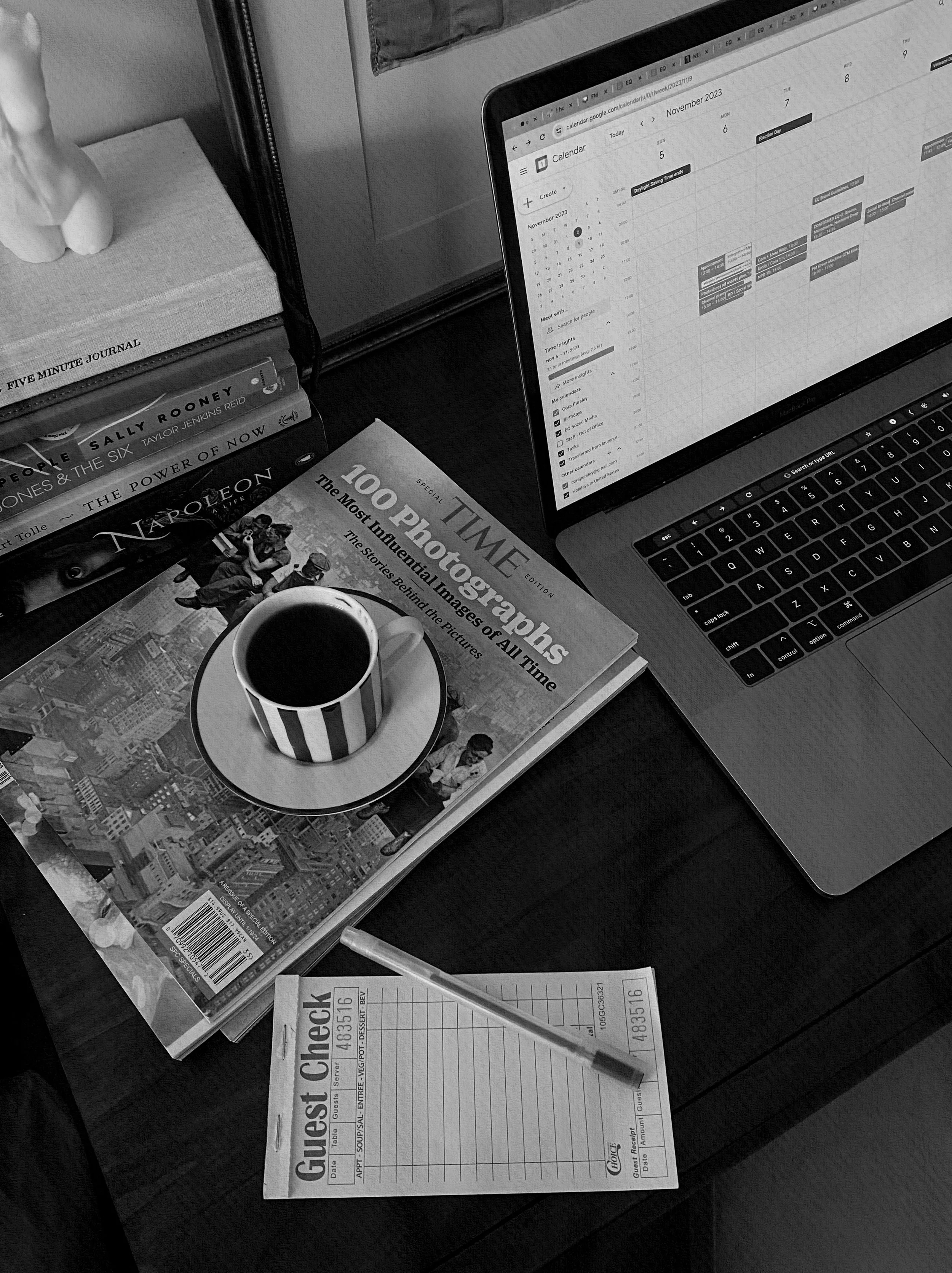 Black and white photo of a workspace with a laptop displaying a calendar, a cup of coffee, a stack of books including a journal and a photography book, and a medical test result form on a dark surface.