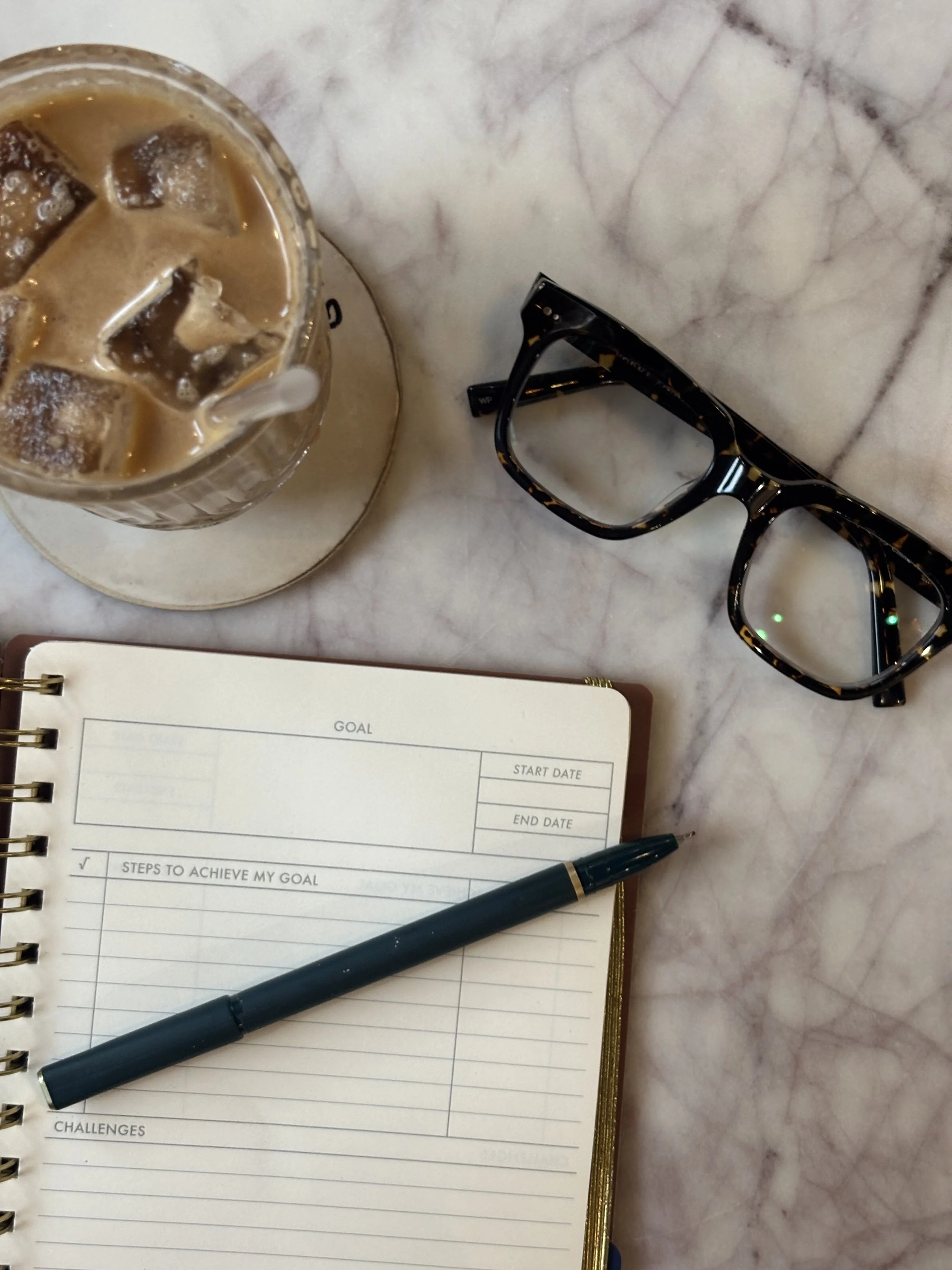 A marble table with a variety of items including a glass of iced coffee, a pair of tortoiseshell glasses, a black pen, and an open planner with pages for setting goals, tracking challenges, and steps to achieve goals.