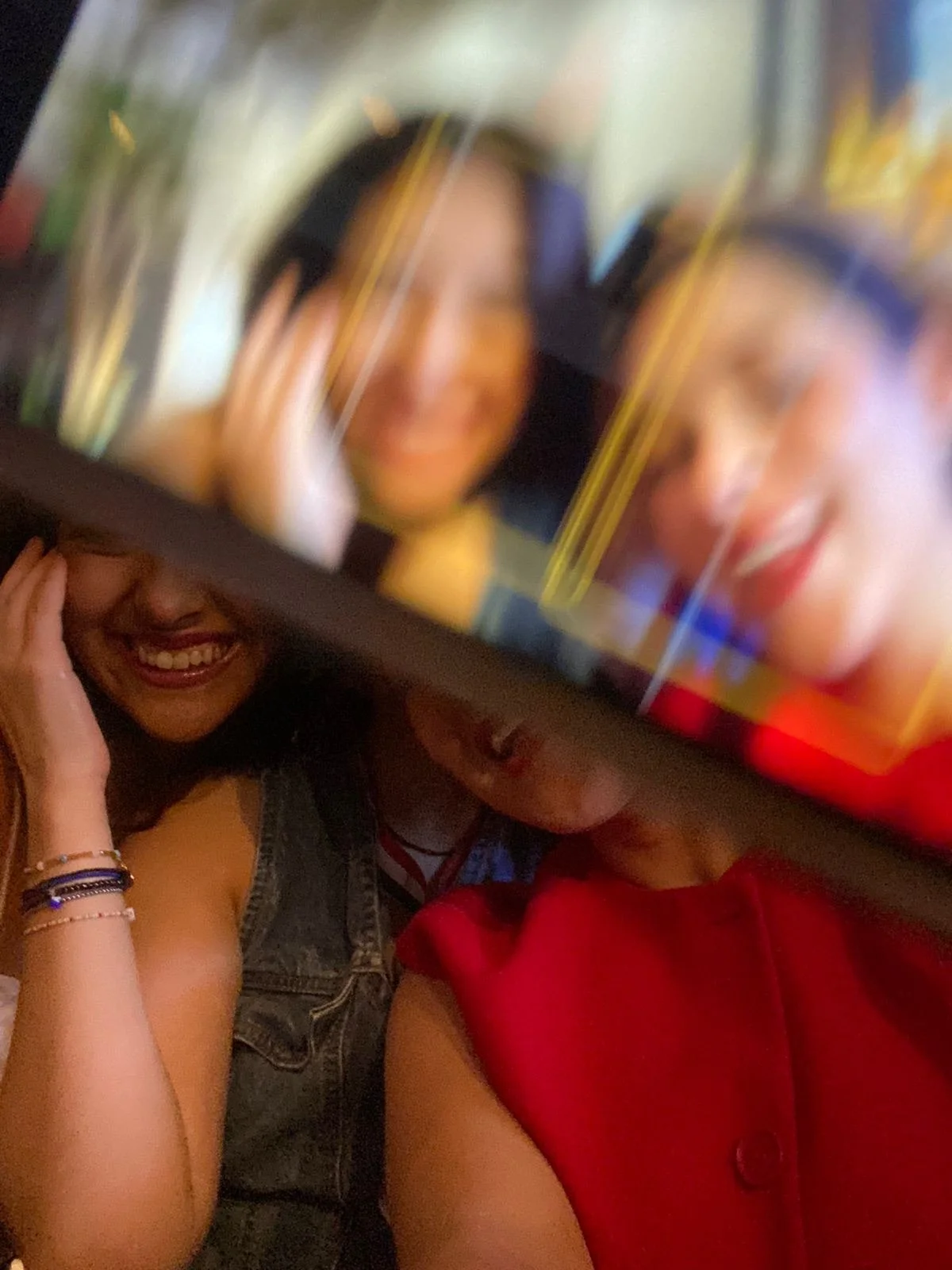Two young women are smiling and appear to be looking at a phone or digital screen. One woman has long dark hair, and the other has long dark hair and glasses. They are close together, with one woman wearing a denim vest and the other a red top, in a lively, colorful setting.