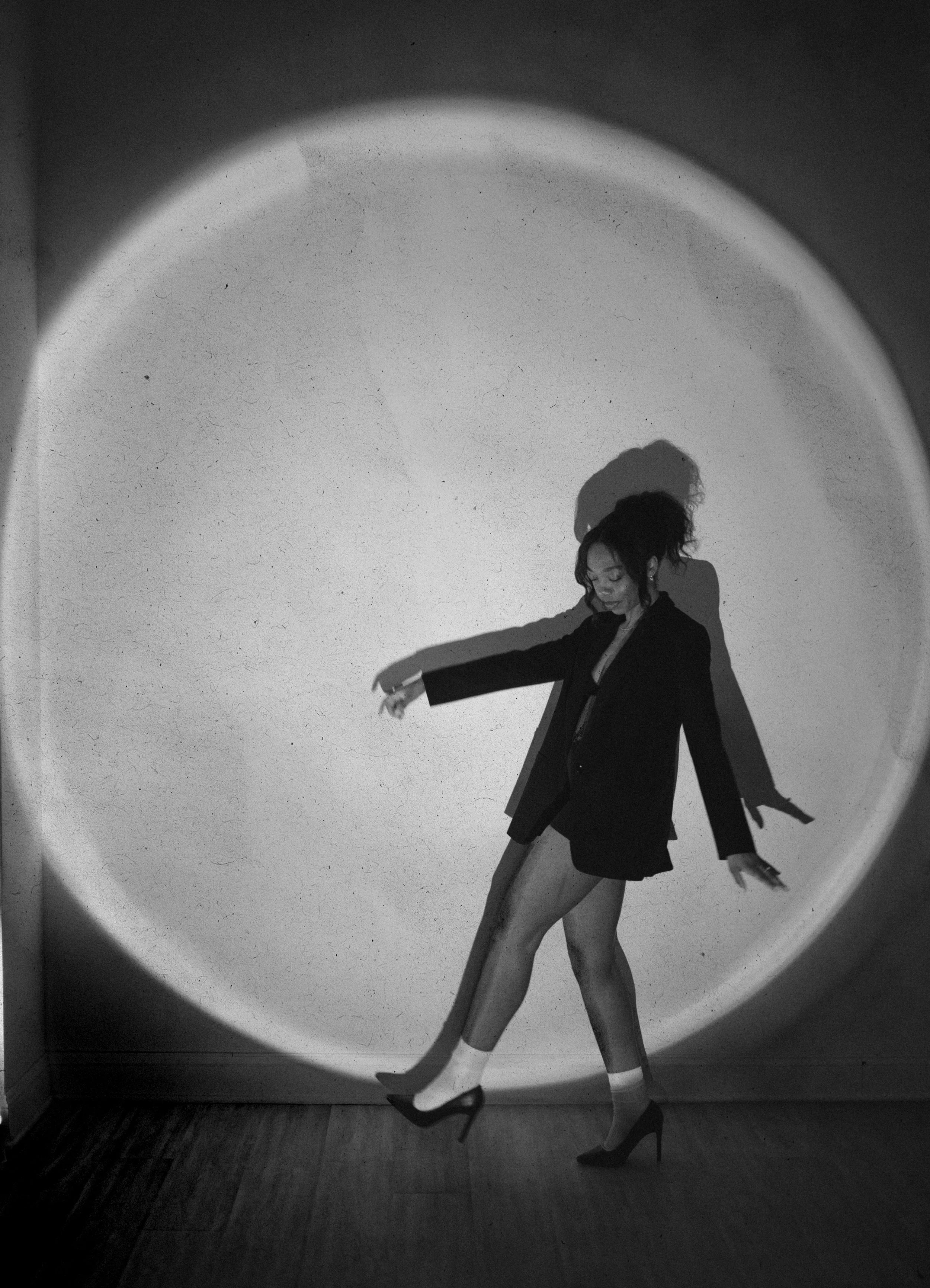 Black and white photo of a woman in high heels, a blazer, and shorts, dancing or posing with her arms extended and her eyes closed, directed downward, near a wall with a circular light or shadow pattern.