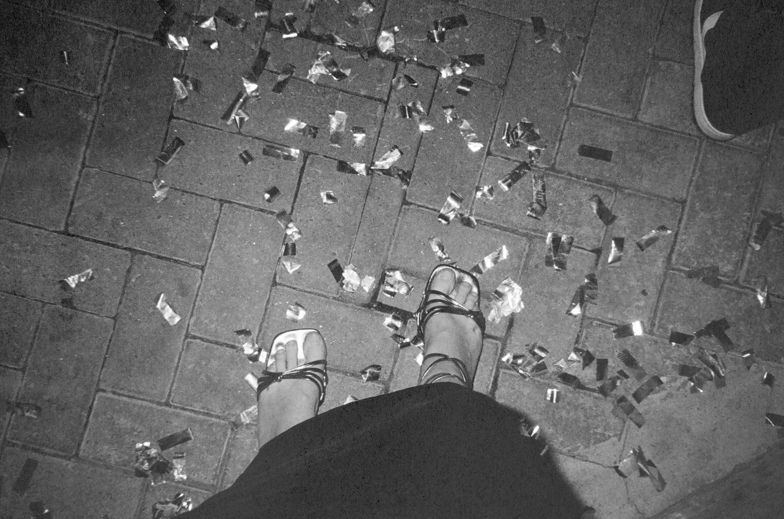 A person's feet in sandals standing on a tiled floor with scattered pieces of crumpled aluminum foil and an adjacent shoe visible in the corner.