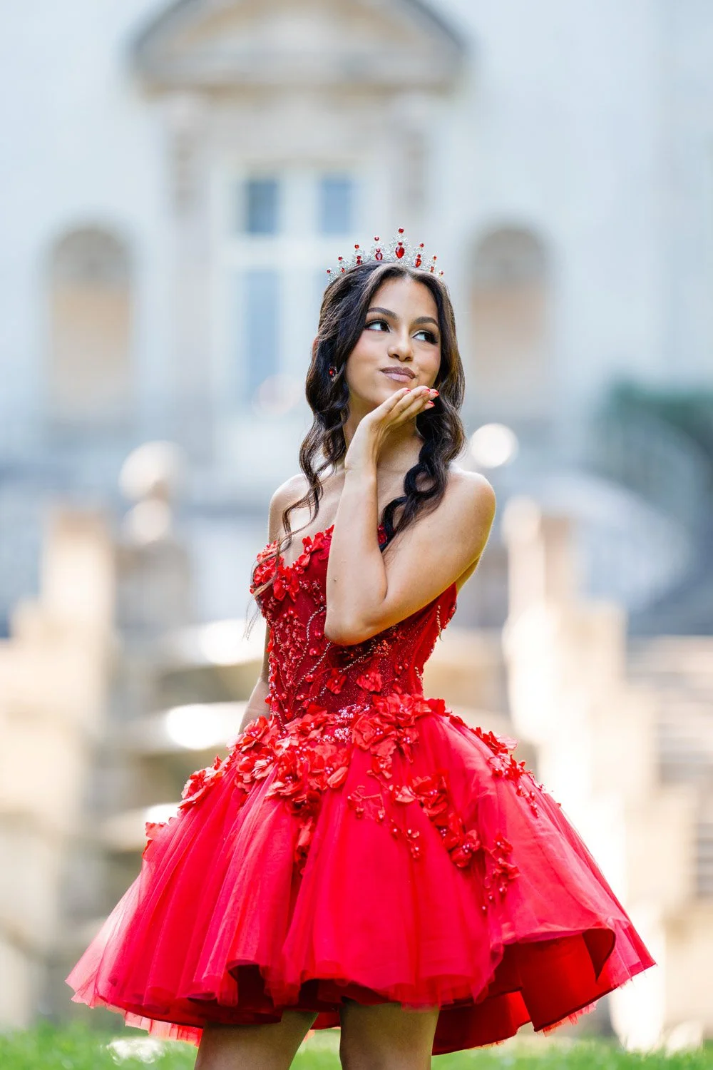 A young woman in a red, floral embroidered dress and tiara, standing outdoors in front of a historic building with fountains, posing with her hand near her chin, making a playful expression.