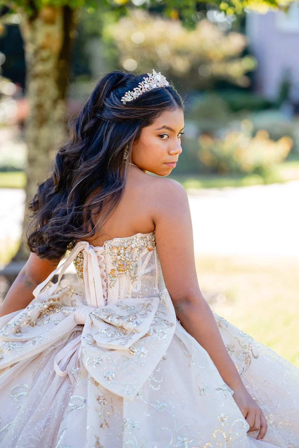 A young woman wearing a light-colored, ornate dress with large bow and embroidery, standing outdoors with trees and greenery in the background, looking over her shoulder.