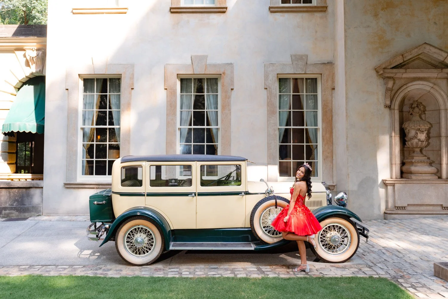 A woman in a red dress and tiara standing next to a vintage beige and green car parked on a cobblestone street in front of a building with large windows and an ornate stone urn sculpture.
