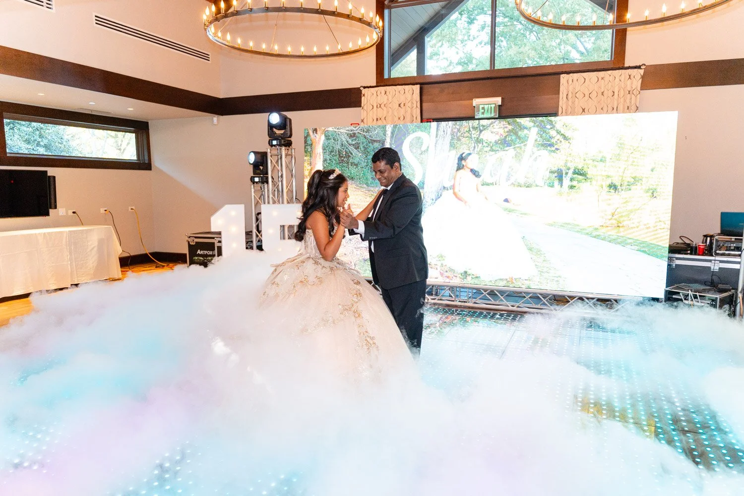 A bride and groom sharing a first dance on a decorated dance floor with smoke effects at a wedding reception hall. The bride is wearing a white wedding gown, and the groom is dressed in a black suit. In the background, there is a large digital screen