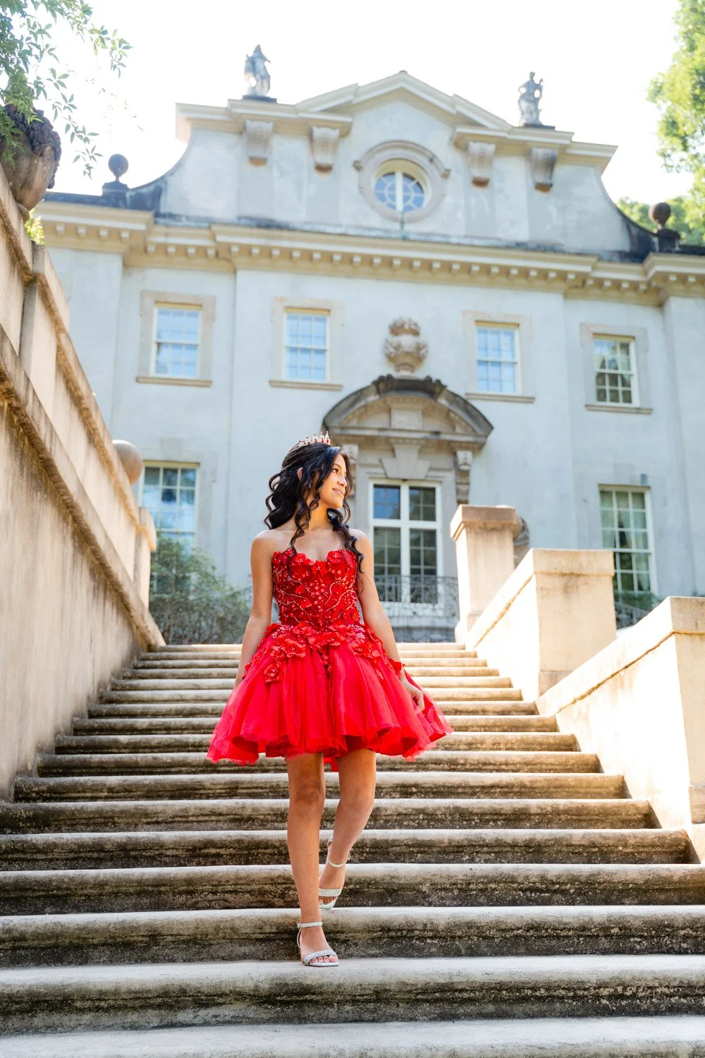 A young woman in a red dress and tiara walking down stone stairs in front of a historic building.