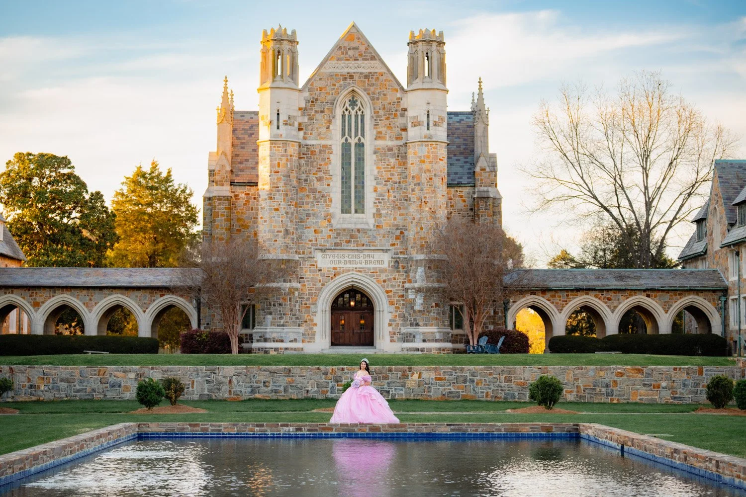A girl in a pink dress standing in front of a stone castle with arched windows and a fountain in the foreground, surrounded by trees and grass during sunset.