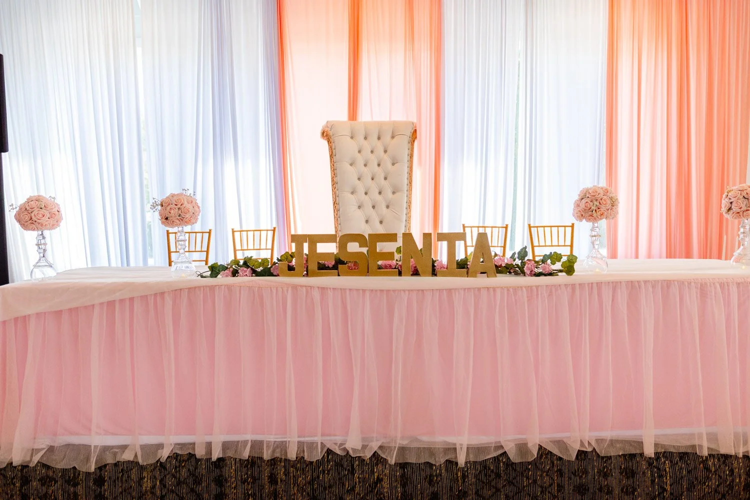 Wedding or event table decorated with pink tulle, floral arrangements with light pink roses, and large gold letters spelling 'JESENTA' in front of a tufted white high-back chair. The background features pastel pink and white curtains.