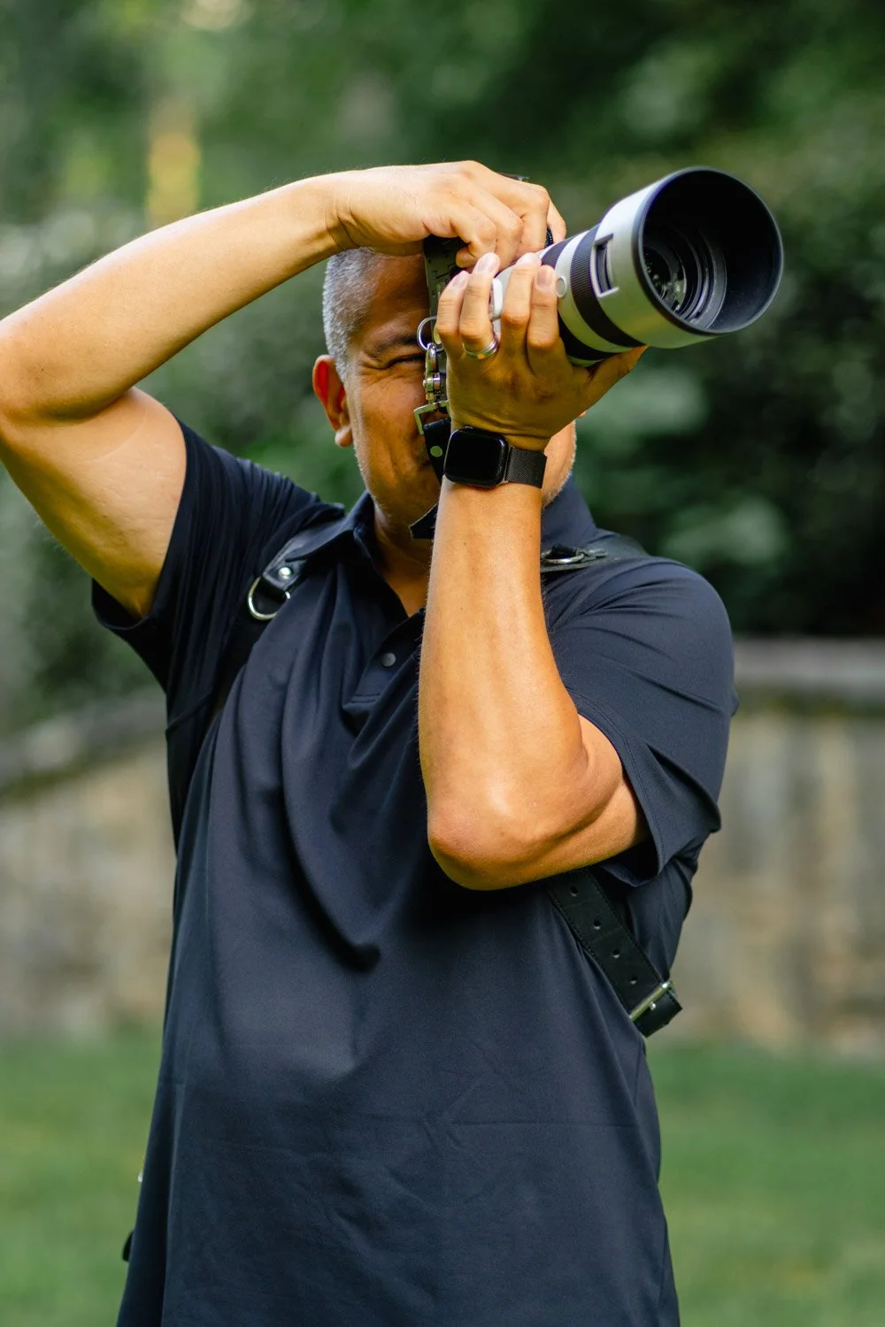 A person with short hair, wearing a black shirt and watch, taking a photograph with a large camera outdoors.