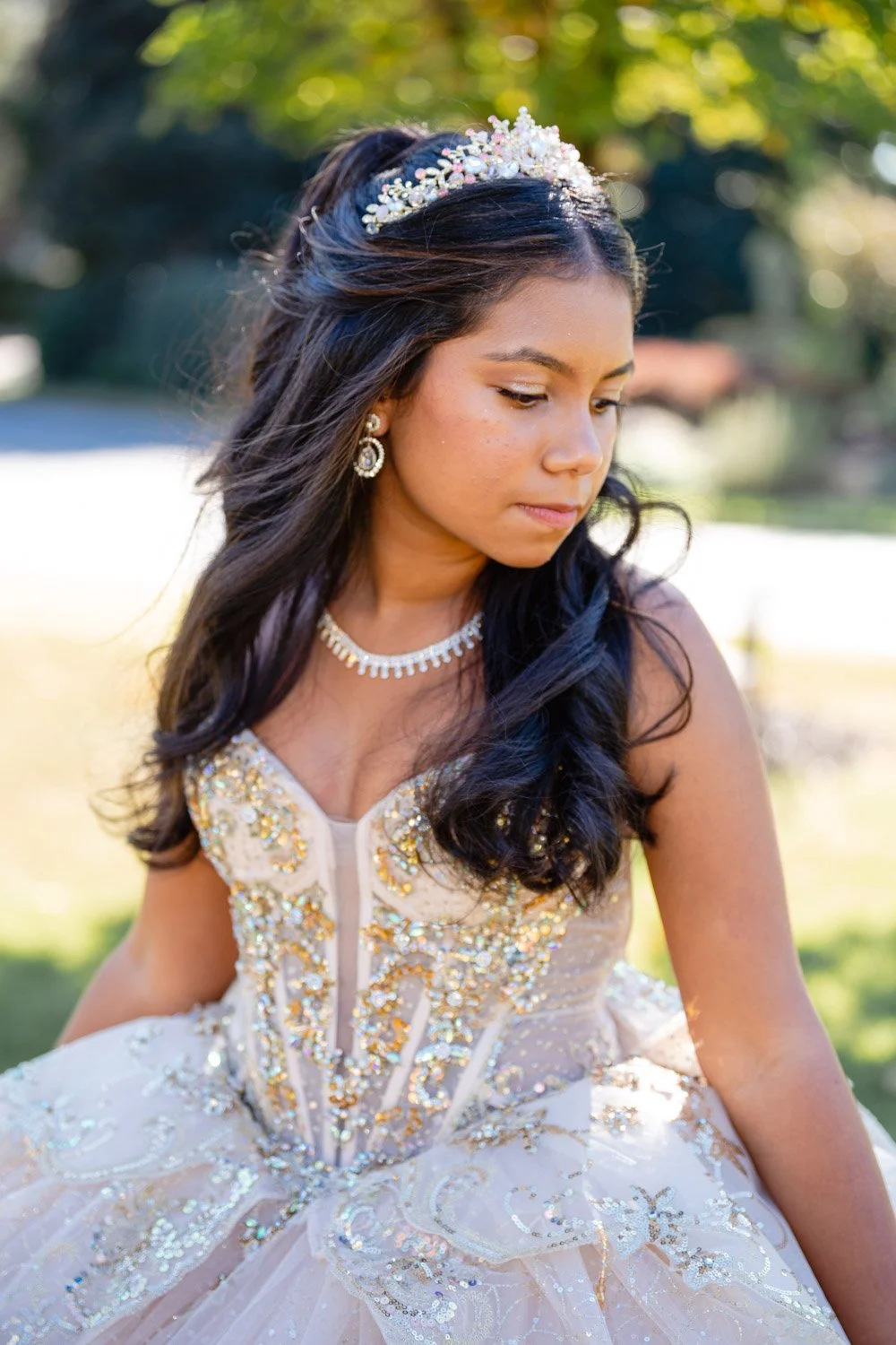 Young woman in a cream-colored, beaded gown with jewelry, wearing a tiara, outdoors with trees in the background.