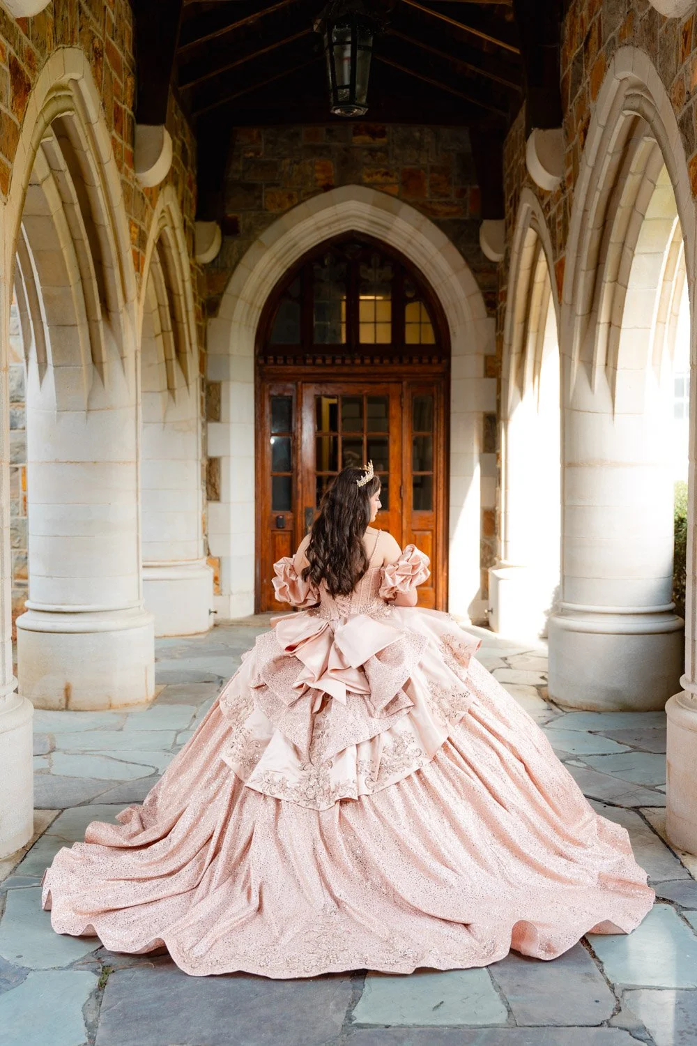 A quinceañera in a pink, ornate ball gown with large bows, lace, and ruffles, standing under a stone archway with Gothic-style windows and doors.