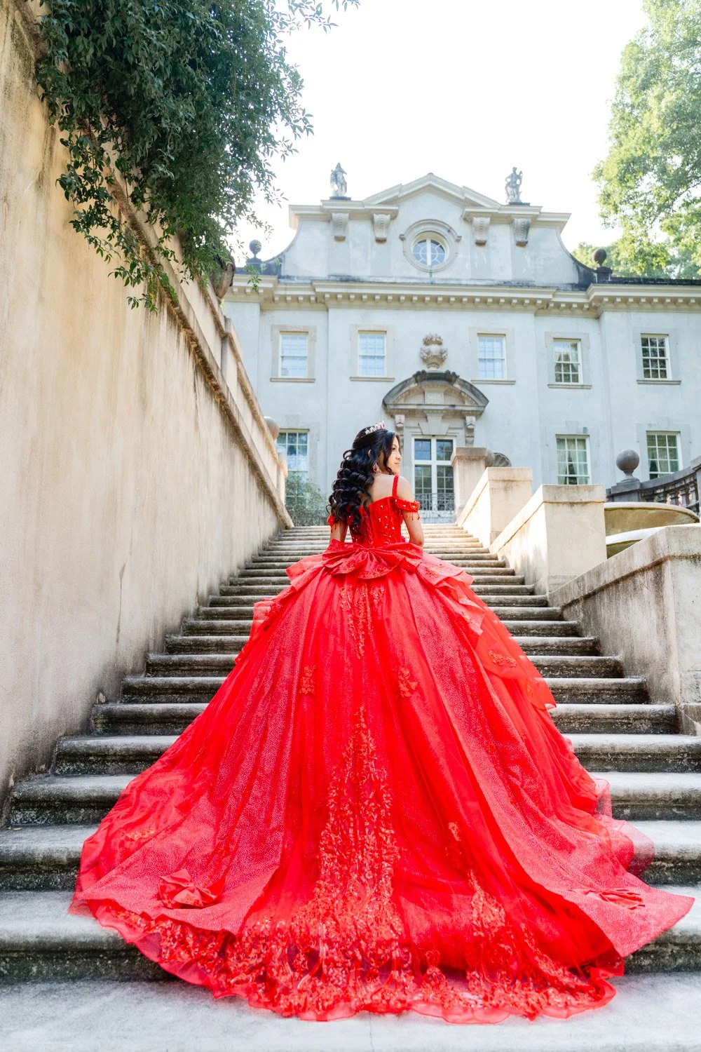 A woman in a red ball gown with an extended train is sitting on stone stairs outside of a historic white mansion with large windows and ornate architectural details, surrounded by greenery.