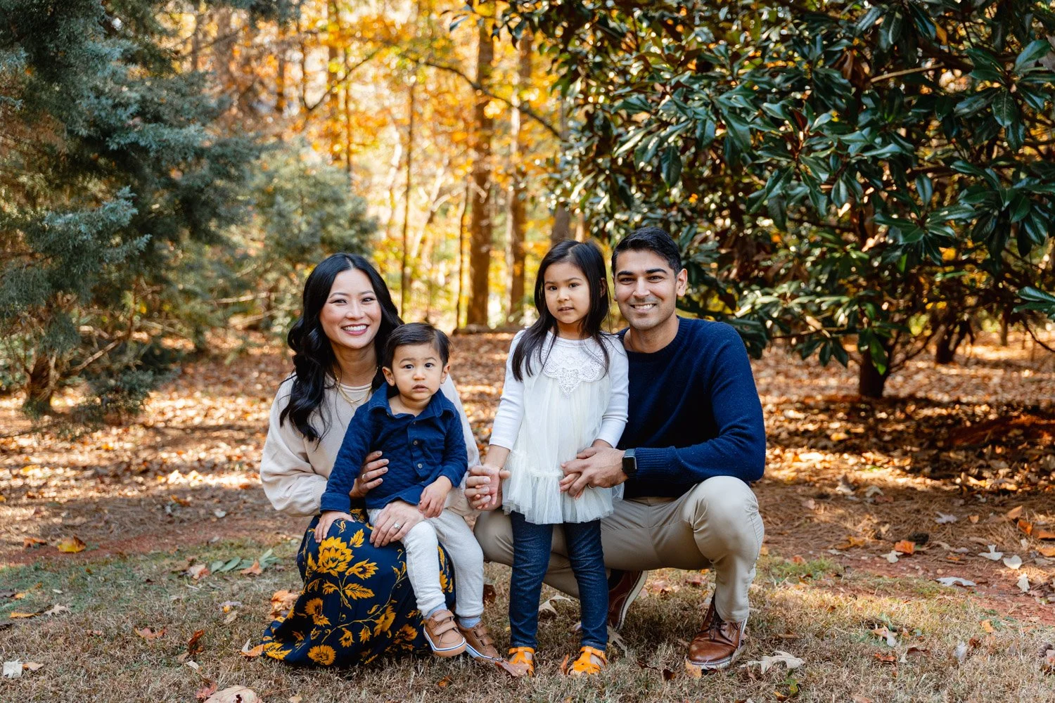 Family of four outdoors in a wooded area with autumn leaves, smiling and posing for a photo.