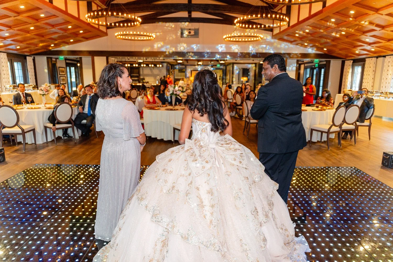 A wedding reception with a bride in a wedding gown standing on a dance floor, accompanied by two women and a man, with guests seated at round tables in the background.