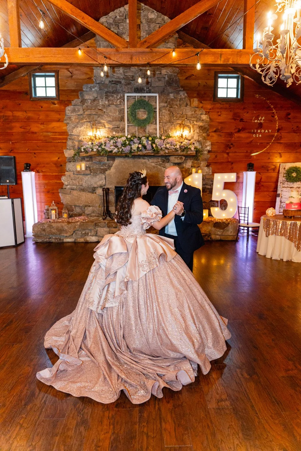 A quinceañera and groom dancing in a rustic wooden wedding venue with a stone fireplace decorated with greenery and flowers, large illuminated number '5' sign, and warm lighting.