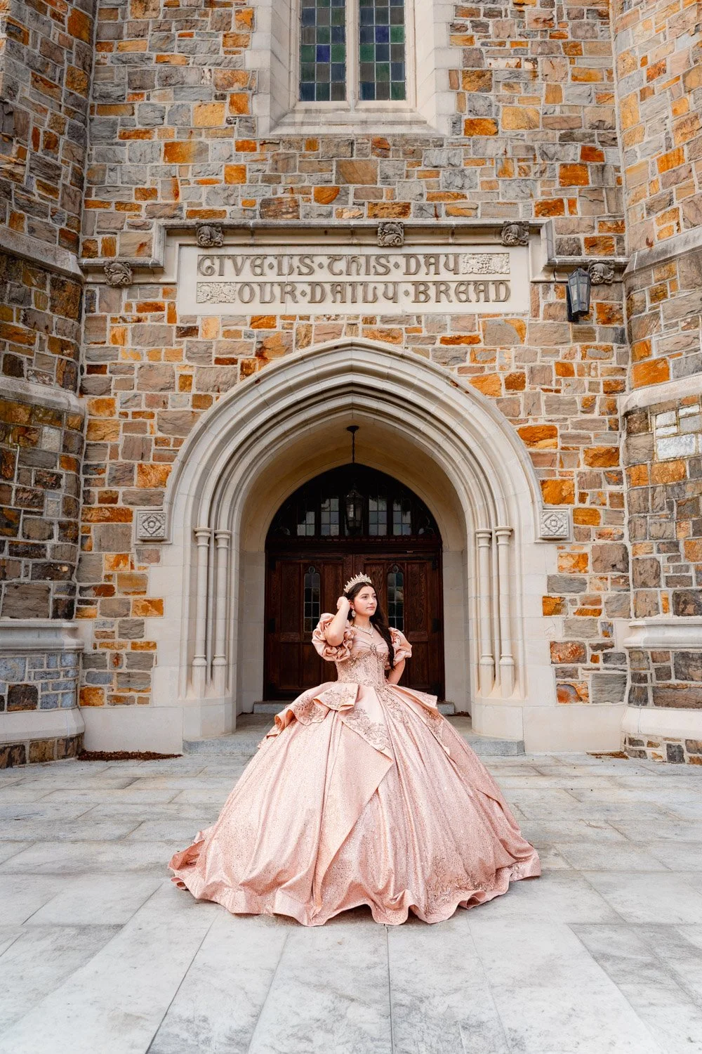 A quinceañera dressed in a peach-colored, elaborate ball gown standing in front of a stone church with arched doorway and stained glass window, wearing a crown.