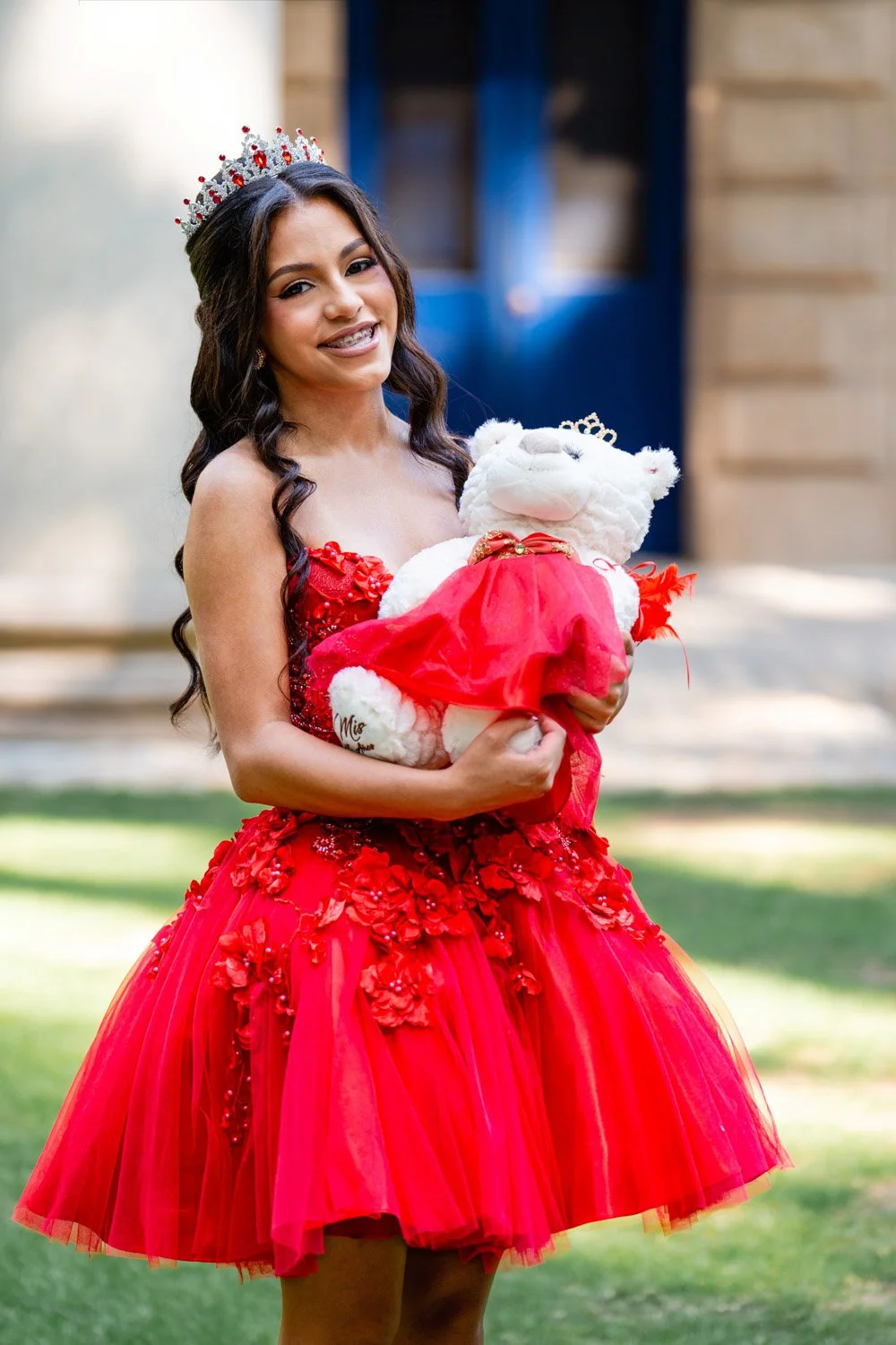 A young woman wearing a red dress and a tiara, holding a white teddy bear dressed in a red dress and a tiara, standing outdoors in front of a building with a blue door.