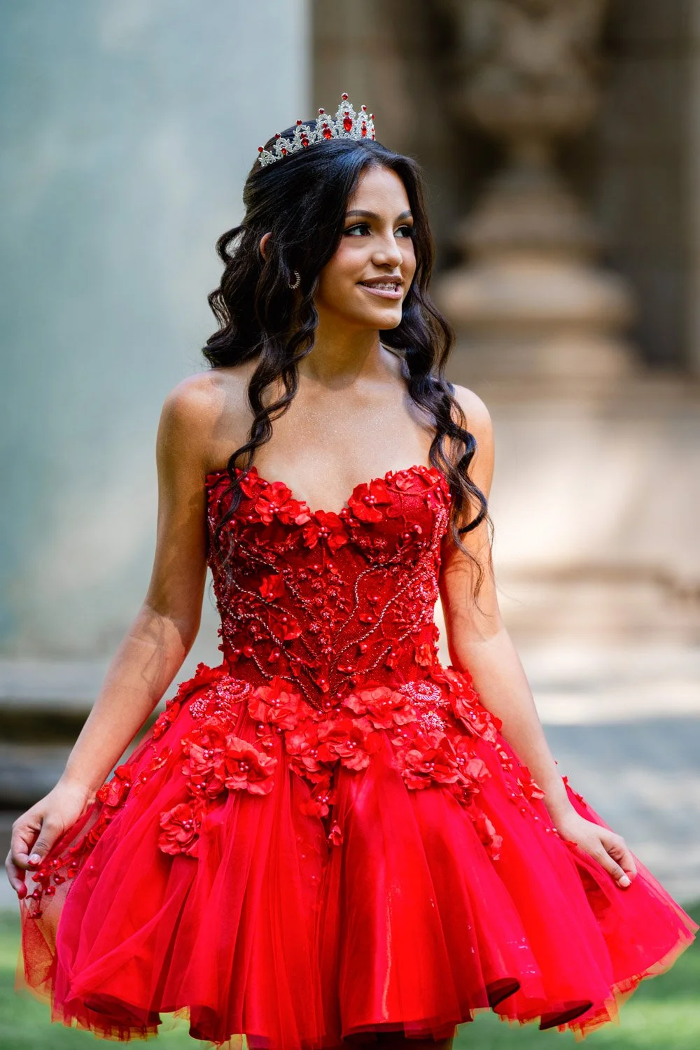 A young woman wearing a red, sleeveless, floral-embellished dress and a silver tiara with red accents, standing outdoors.