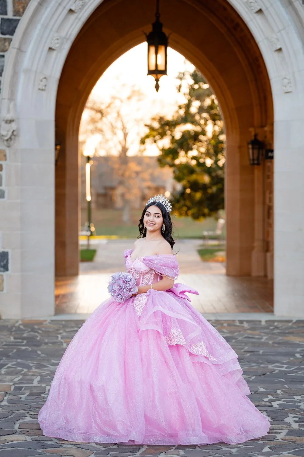 A young woman in a pink ball gown with off-the-shoulder sleeves and a full skirt, wearing a tiara and holding a bouquet of lavender roses, stands under an arched stone gateway with lanterns, outdoor setting at sunset.