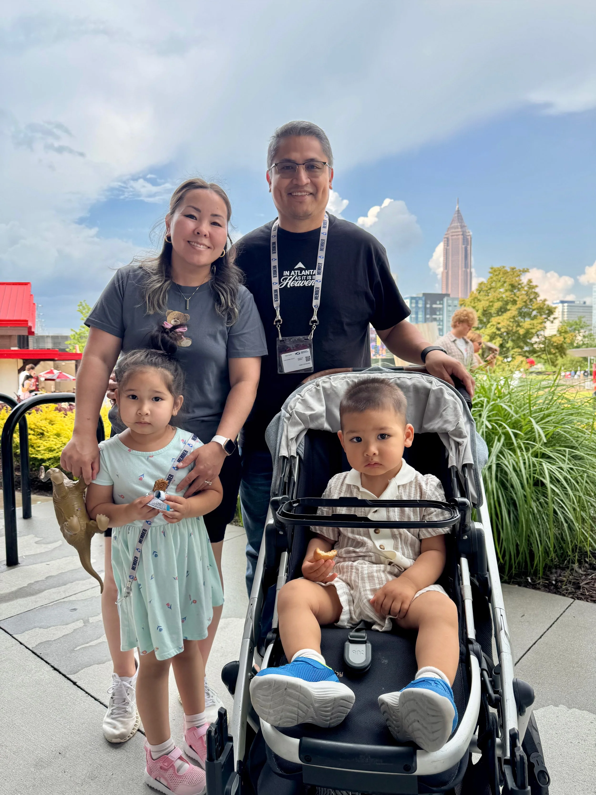 A family of five posing outdoors with the Atlanta skyline in the background. The family includes a woman, a man, a young girl, and two young boys, one in a stroller. The woman is wearing a gray t-shirt and the man is wearing a black t-shirt with a lanyard.