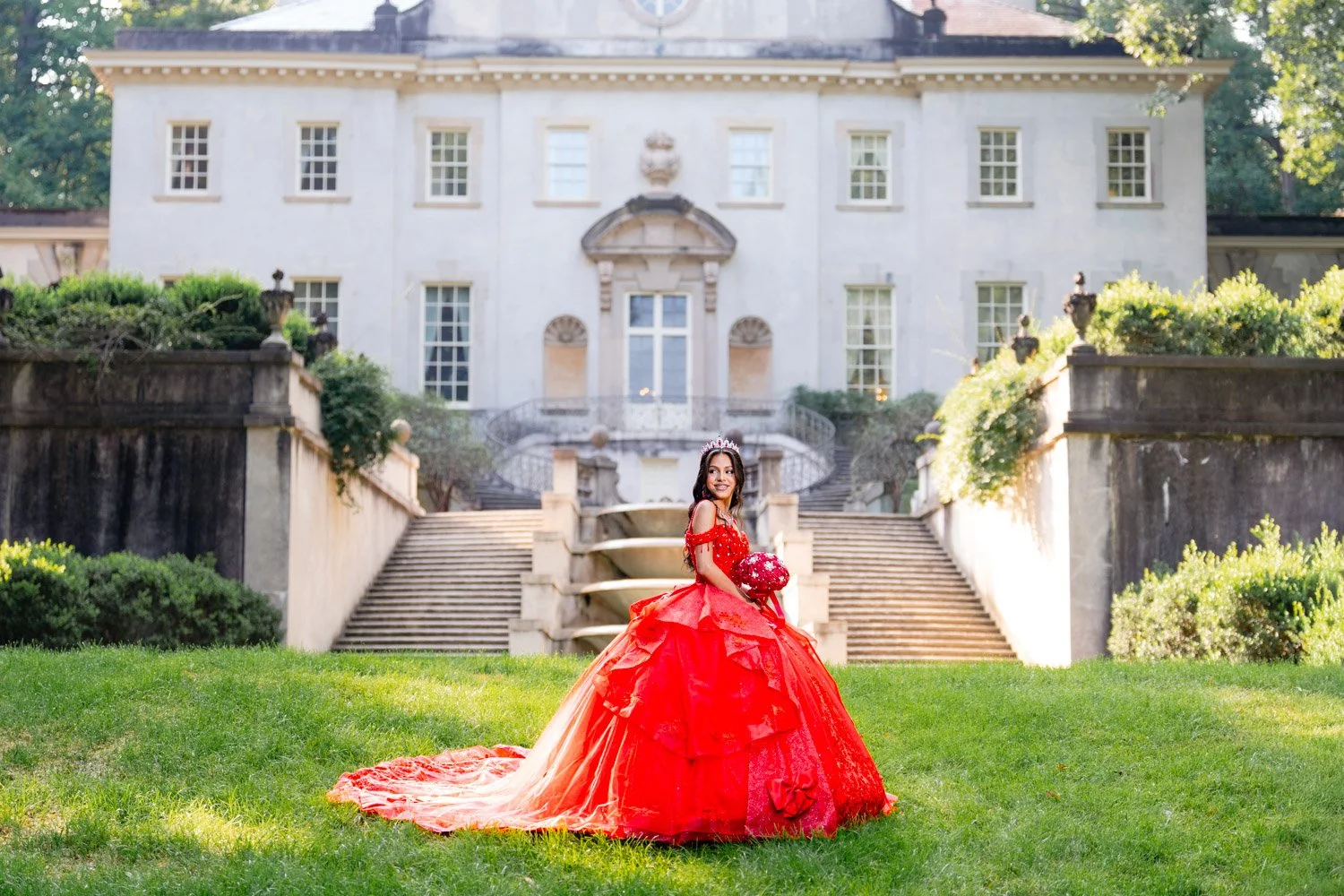 A quinceañera in a red ball gown with a tiara holding a bouquet standing on a grassy lawn in front of a grand white mansion with stairs and greenery.