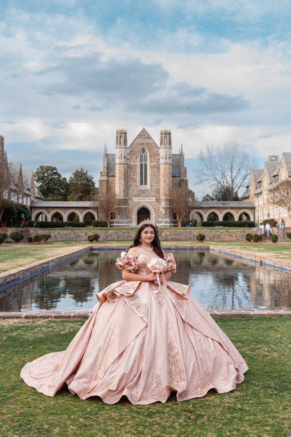 A quinceañera in a pink ball gown and tiara stands by a rectangular pond in front of a historic stone church, holding a bouquet of flowers.