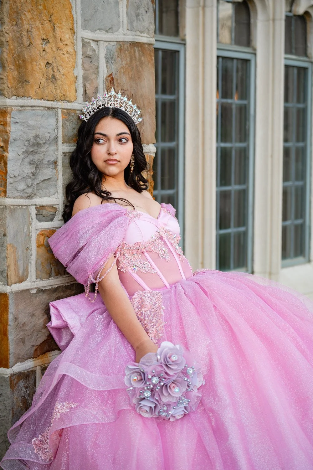 A young woman in a pink princess dress with a tiara and holding a bouquet of pink paper roses, standing against a stone wall with windows in the background.