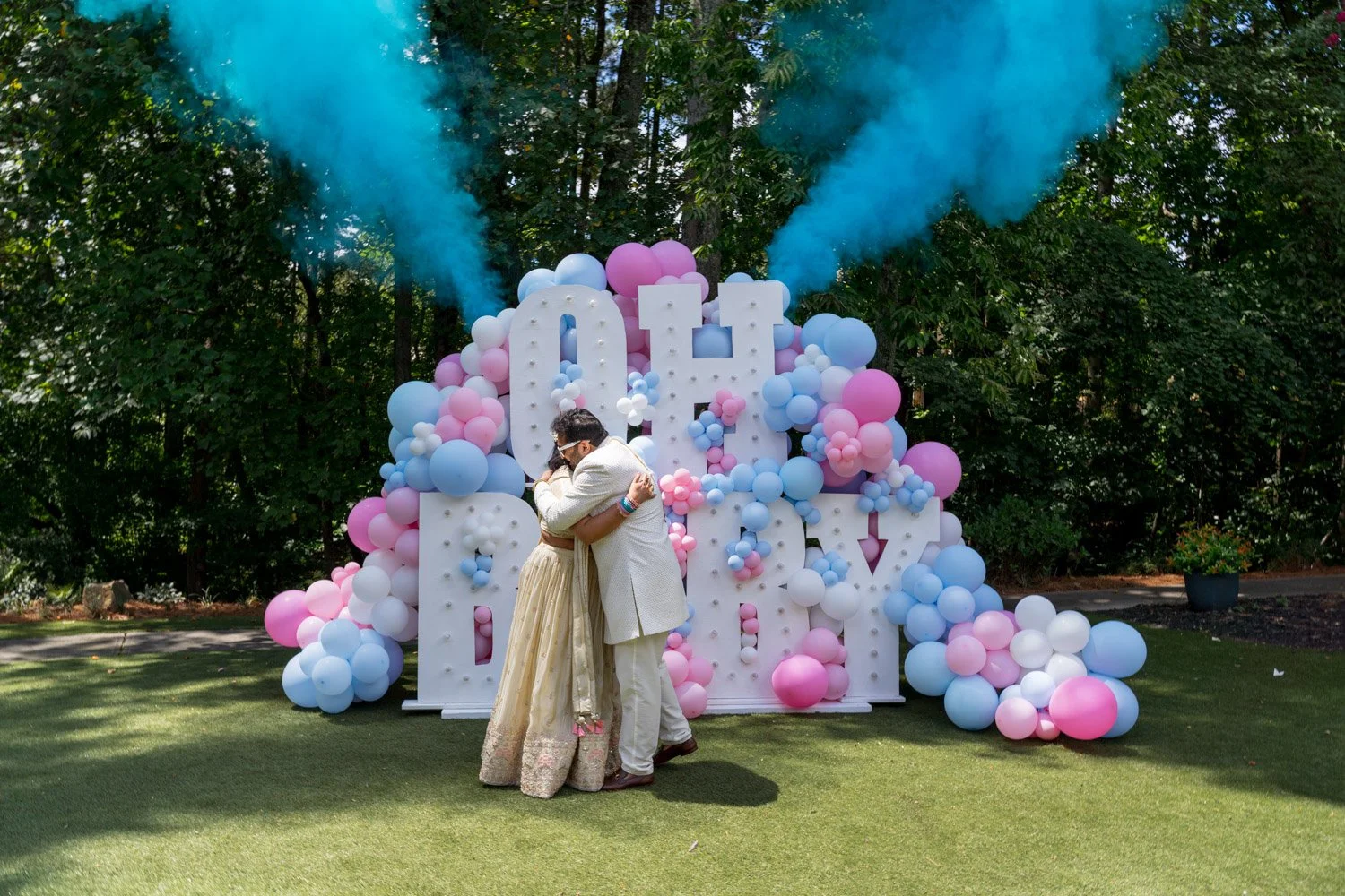 A couple hugging at their wedding, standing in front of a decorative backdrop with large white letters spelling 'LOVE' surrounded by pink, blue, and white balloons with blue smoke clouds overhead.
