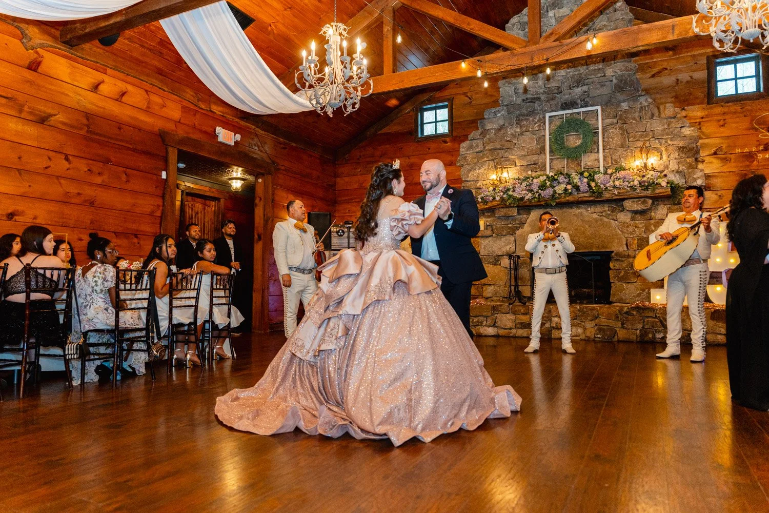 A bride and groom dance in a rustic wedding venue with wood-paneled walls, a stone fireplace, and string lights. The bride wears a voluminous, sparkling ball gown, and the groom is in a dark suit. Wedding guests and musicians are present, with some guests seated at tables and a mariachi band performing near the fireplace.