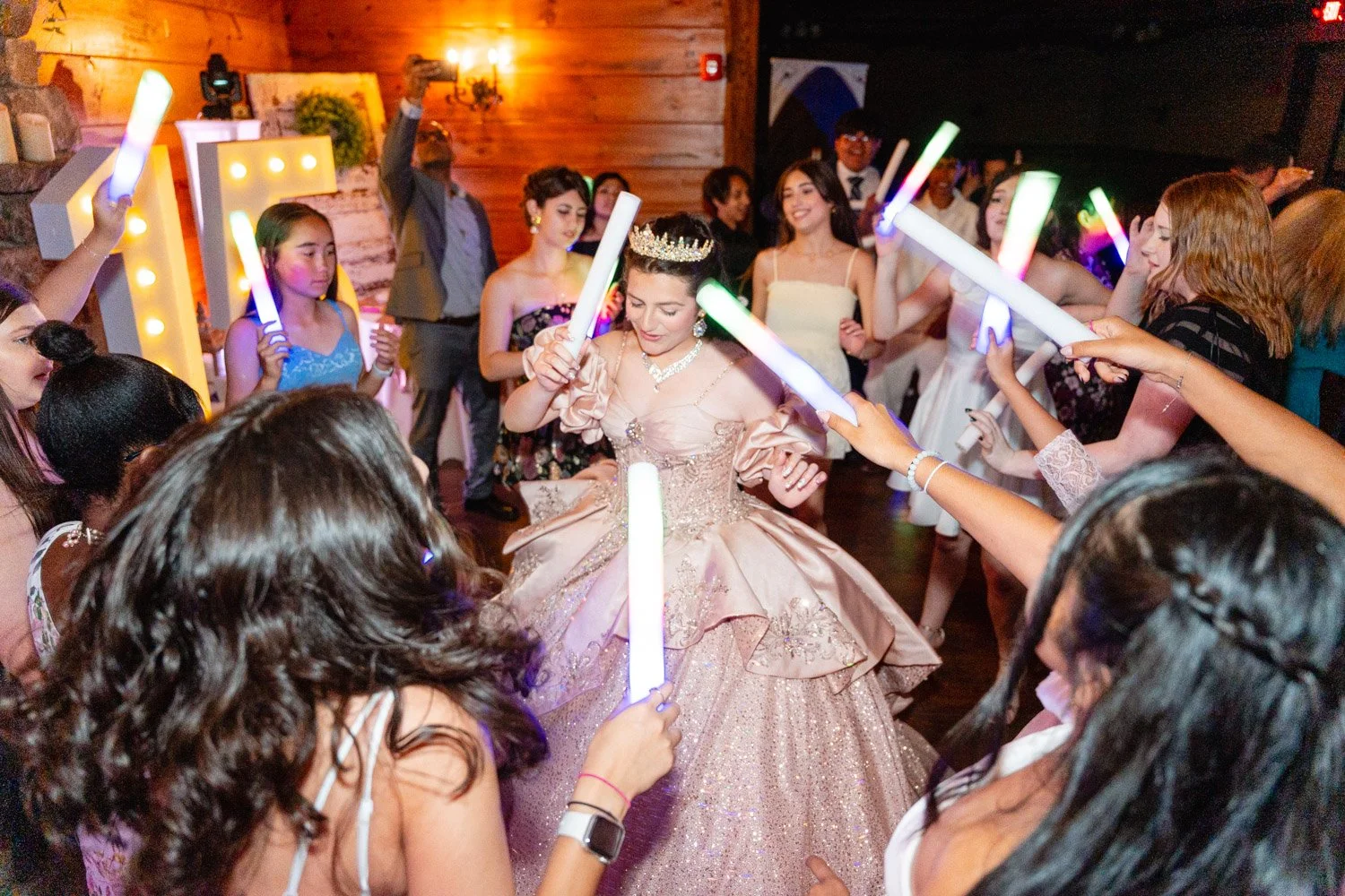 A young woman in a pink gown and tiara dances with friends holding glowing sticks at her sweet sixteen celebration in a decorated indoor venue.