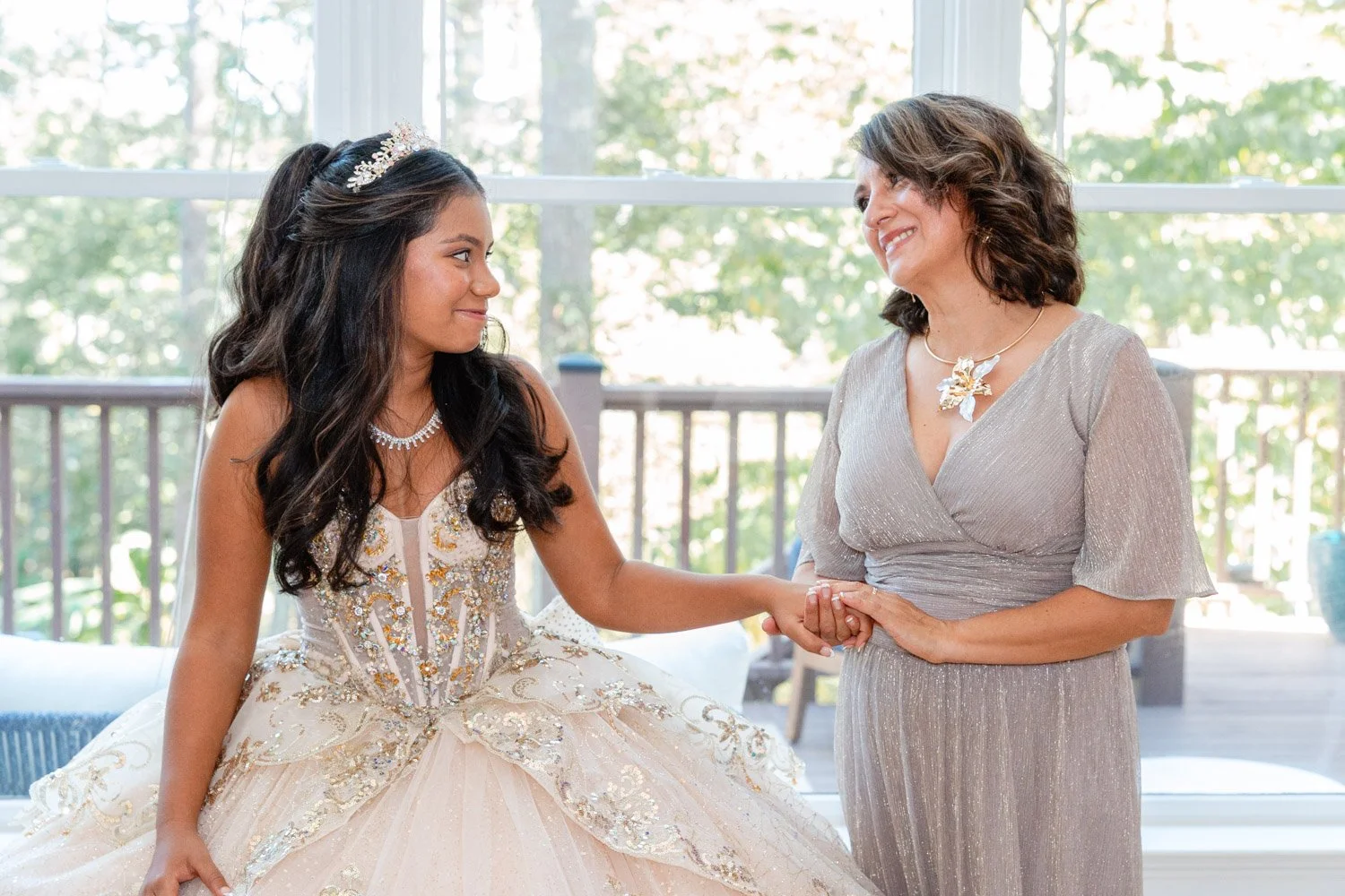 A young girl in a fancy dress and tiara holding hands with an older woman in a silver dress, both smiling at each other inside a bright room with large windows and a deck outside.