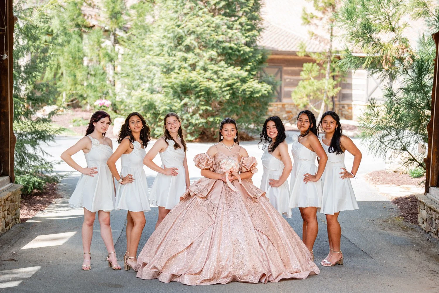 A group of seven young women, one in a pink quinceañera gown holding a bouquet, and six in white dresses, posing under a wooden archway with greenery, trees, and a rustic building in the background.