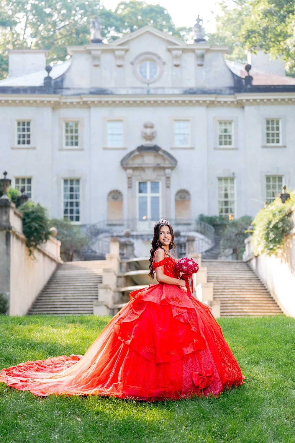 Young woman in a red formal gown holding a bouquet of red flowers, standing on a lawn in front of a grand white mansion with stairs and large windows.
