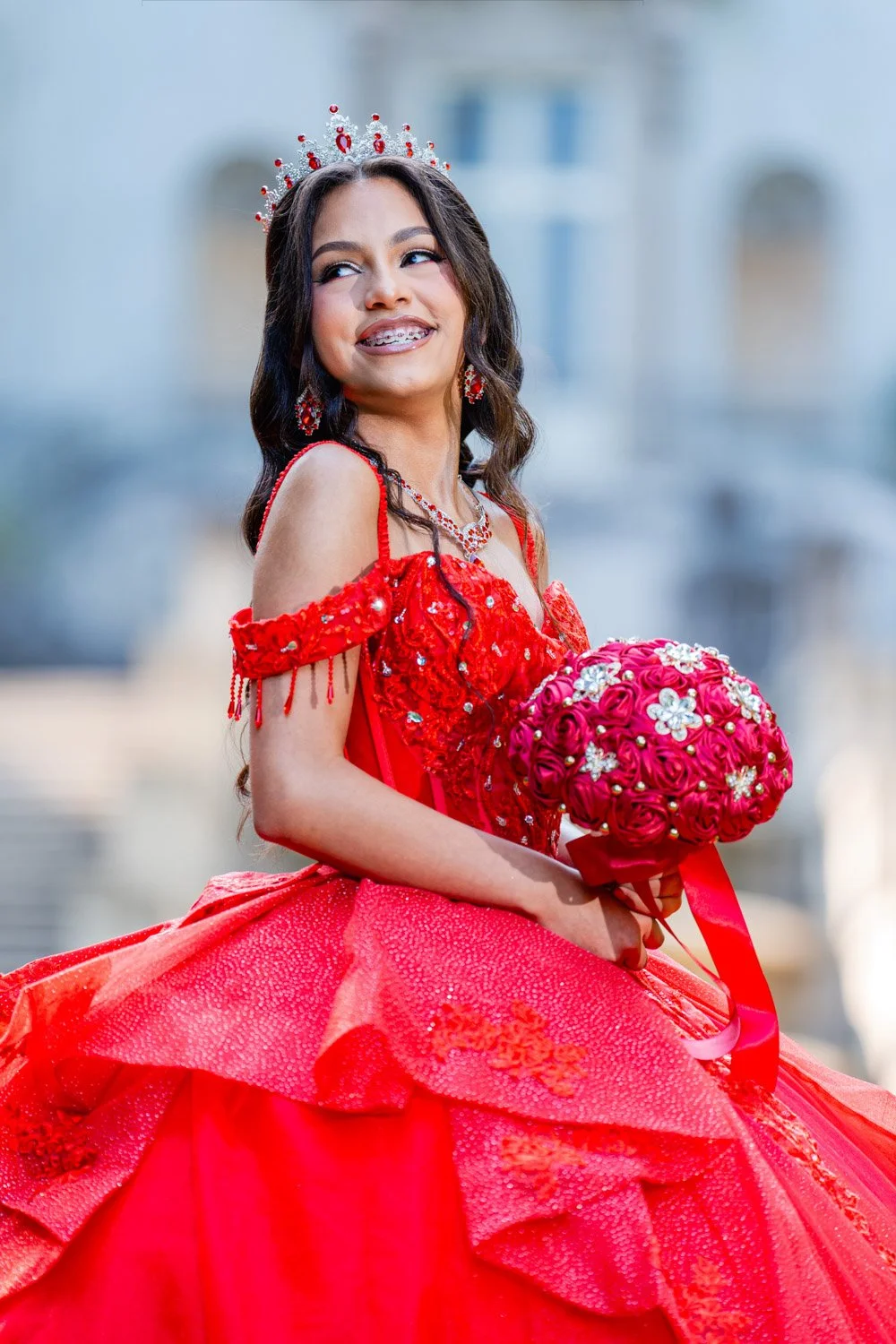 Young woman in a red dress with a tiara, holding a bouquet of pink roses, smiling outdoors.