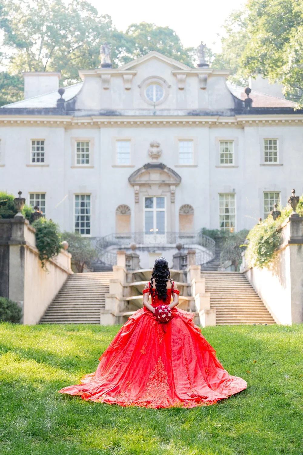 A woman in a red gown holding a bouquet of flowers faces a large mansion with stairs leading up to its entrance, surrounded by greenery.