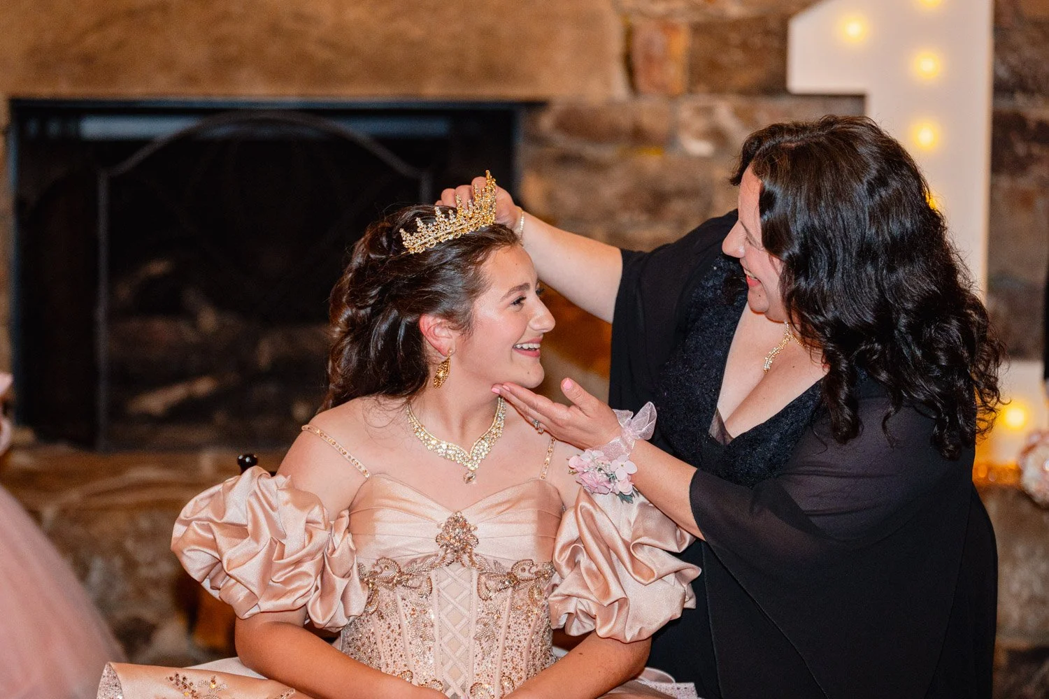 A woman is crowning a young girl dressed in a princess gown with a tiara, suggesting a ceremony like a quinceañera or sweet sixteen celebration.