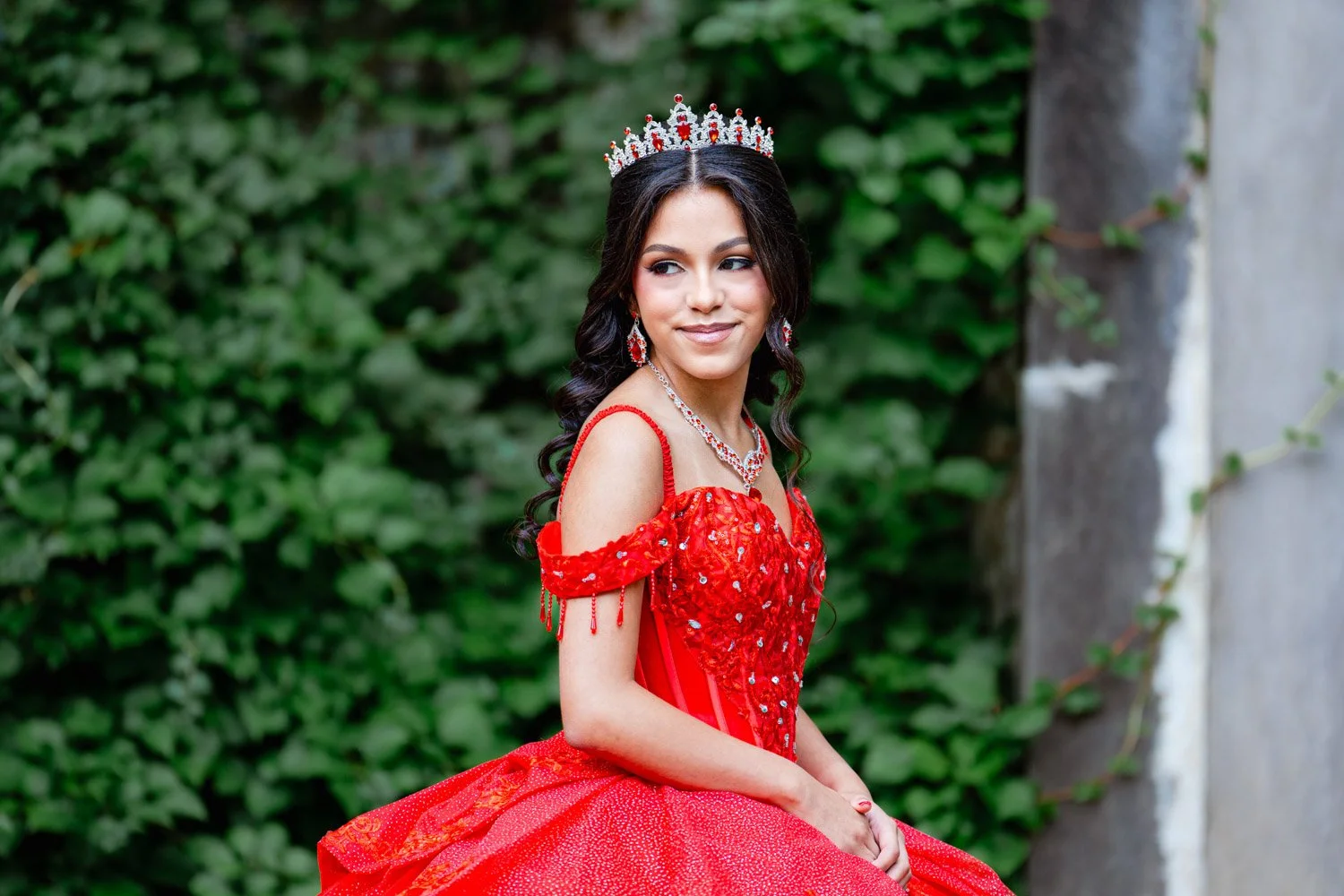 Quinceañera wearing a red gown and tiara against a garden backdrop
