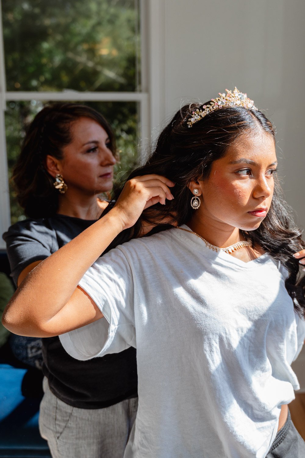 A young woman with long dark hair wearing a tiara, diamond earrings, and a white shirt, getting ready as another woman with shoulder-length dark hair, earrings, and a black top adjusts her hair inside a room with a window and greenery outside.