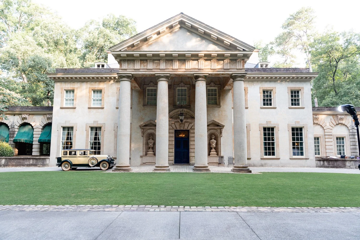 A large, historic mansion with a grand front entrance featuring six columns, surrounded by lush green trees. An antique car is parked on the lawn in front of the mansion.